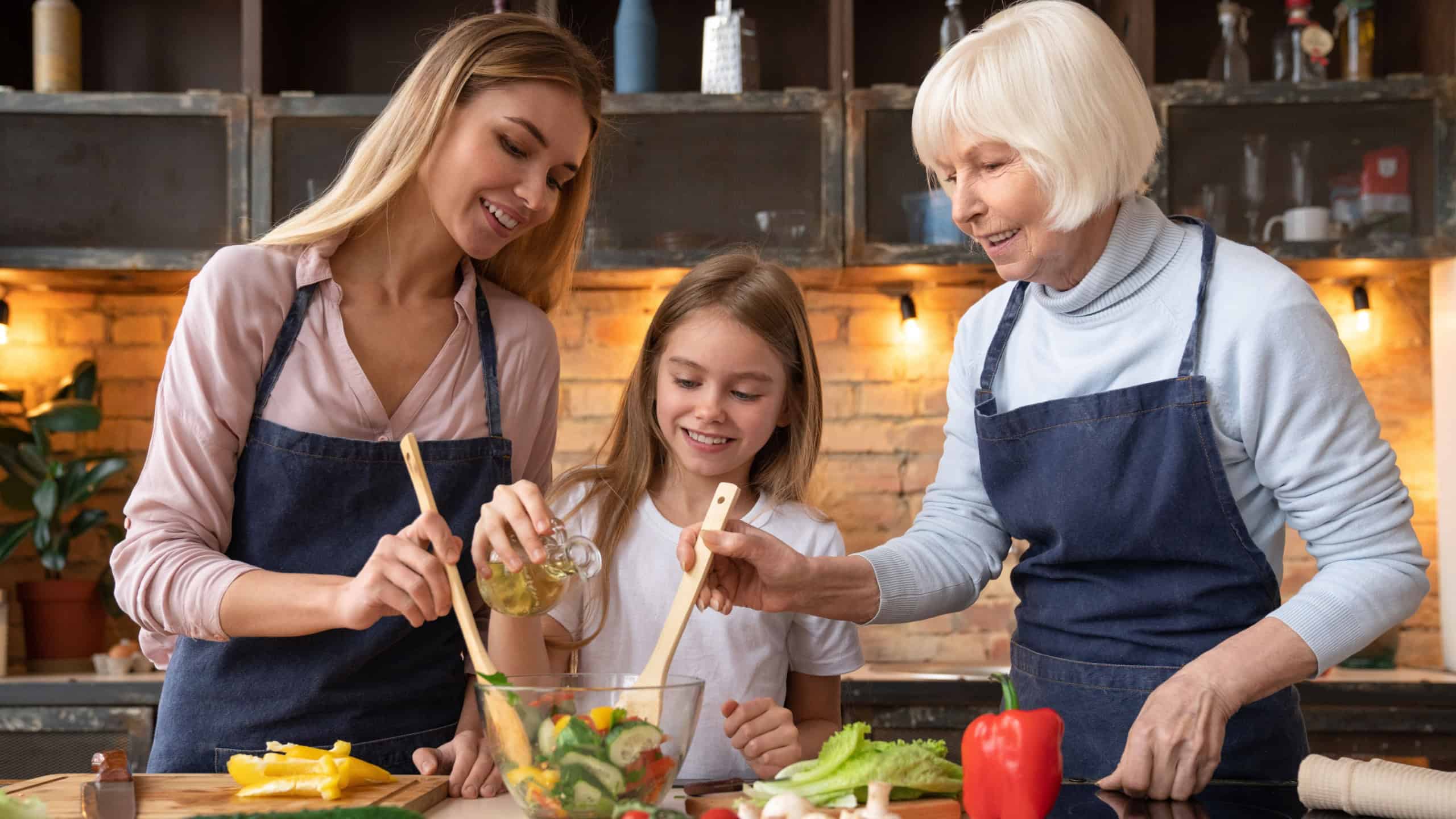 Front view of beautiful little girl that pours oil in salad while young mother and grandmother helping her to mix it. Three generations of women cooking healthy food at home