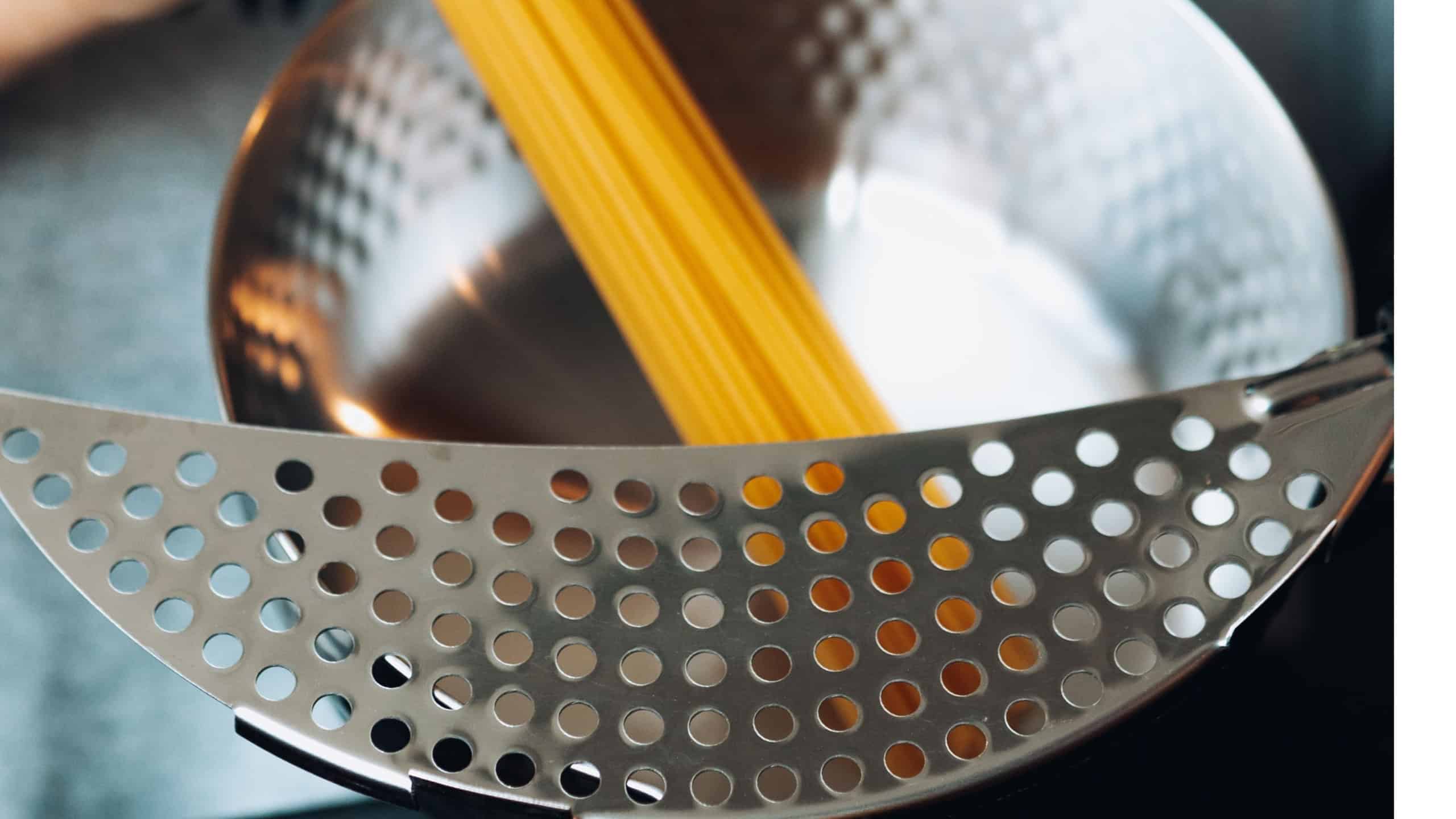 A professional chef stirring al dente pasta in a metal strainer before transferring it to a white ceramic bowl