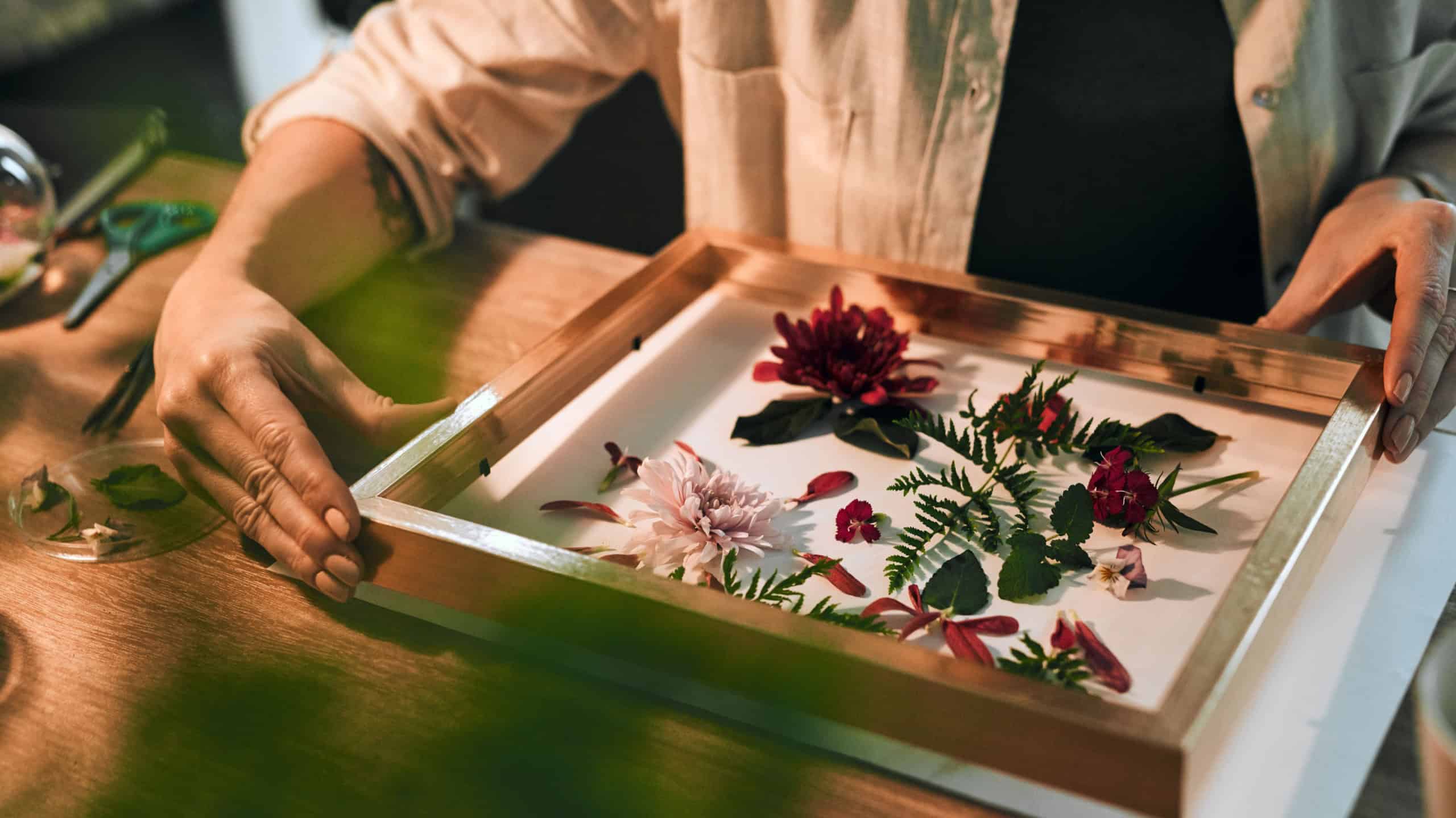 Person, hands and flowers in framing shop with sustainability work or floral, job or decoration on table. Business owner, plants and frame on desk in store with leaf, art design with wood for retail