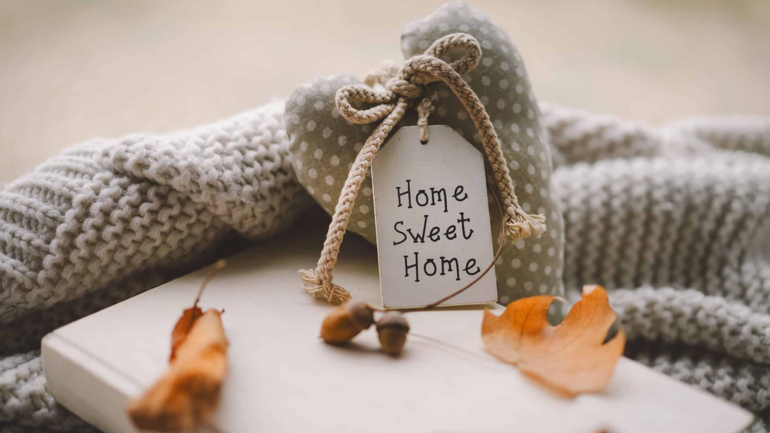 A heart-shaped decorative piece with a tag that says Home Sweet Home is placed on a knitted blanket, surrounded by fallen autumn leaves, creating a warm, inviting atmosphere.