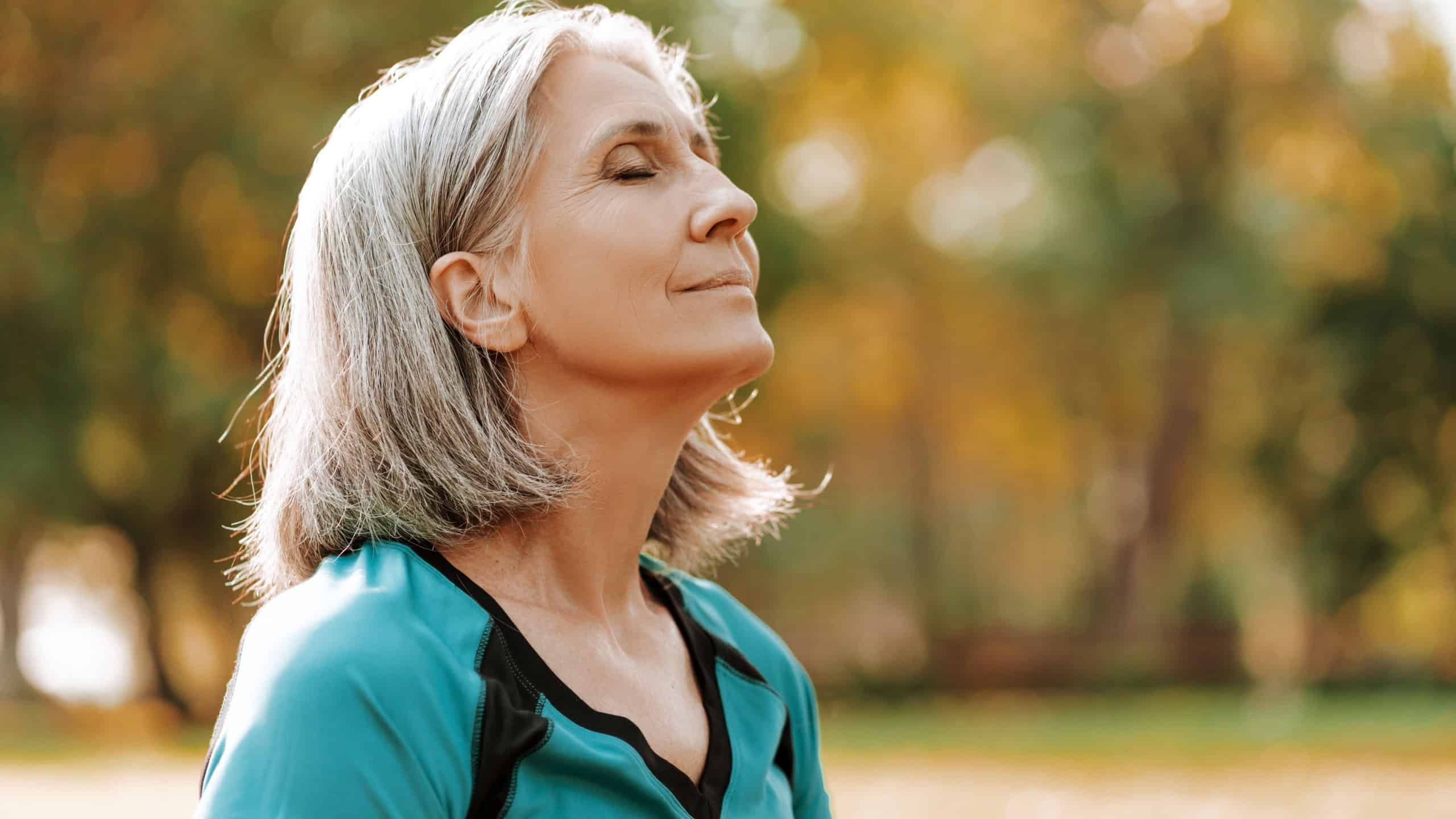 Senior woman wearing sportswear is enjoying a sunny autumn day in the park, closing her eyes and taking a deep breath of fresh air. Balance, harmony concept