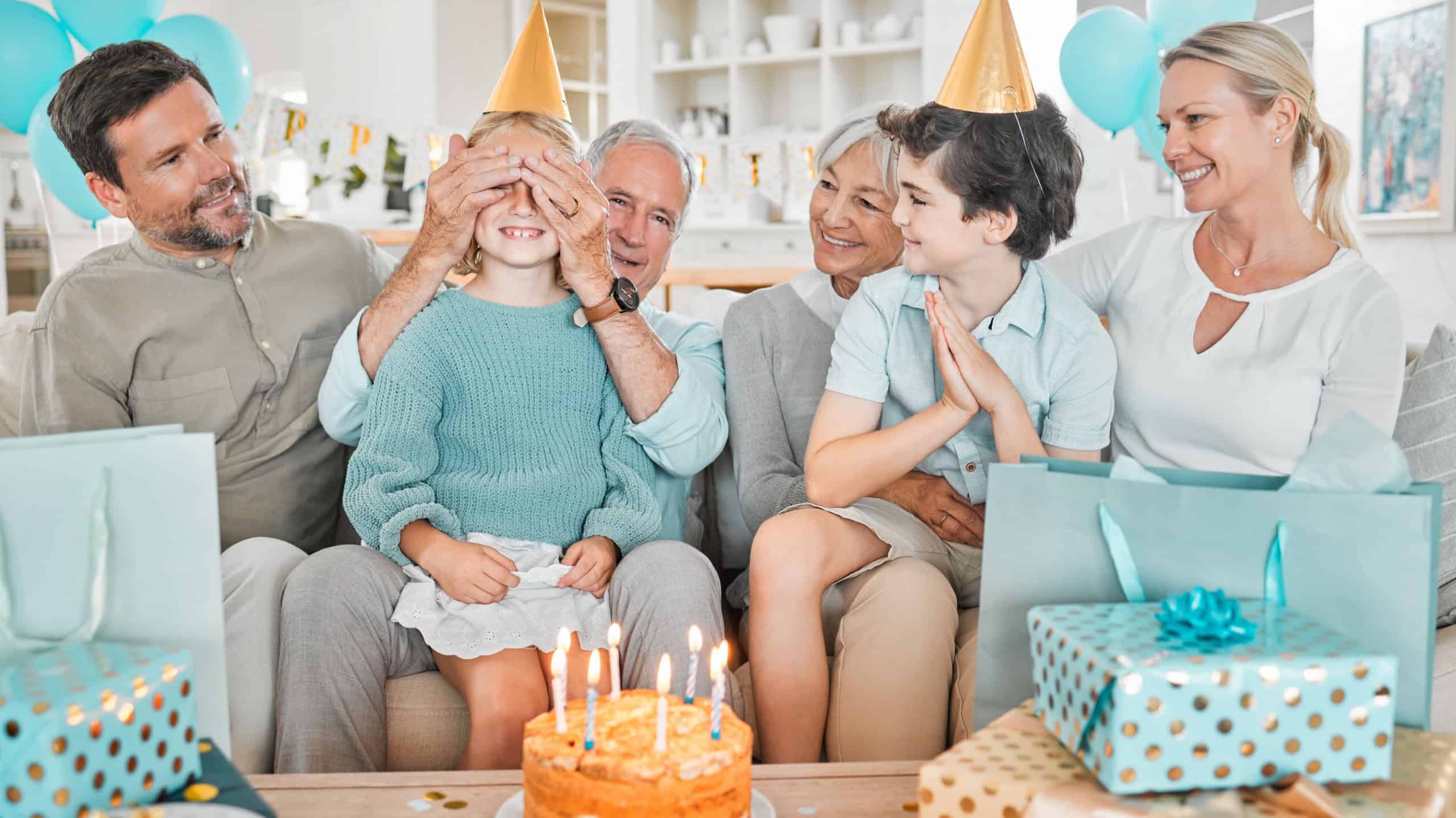 Parents, children and grandparents on sofa for birthday party with cake, games and blindfold for surprise in house. People, playful and kids on couch with present, excited and dessert at family home