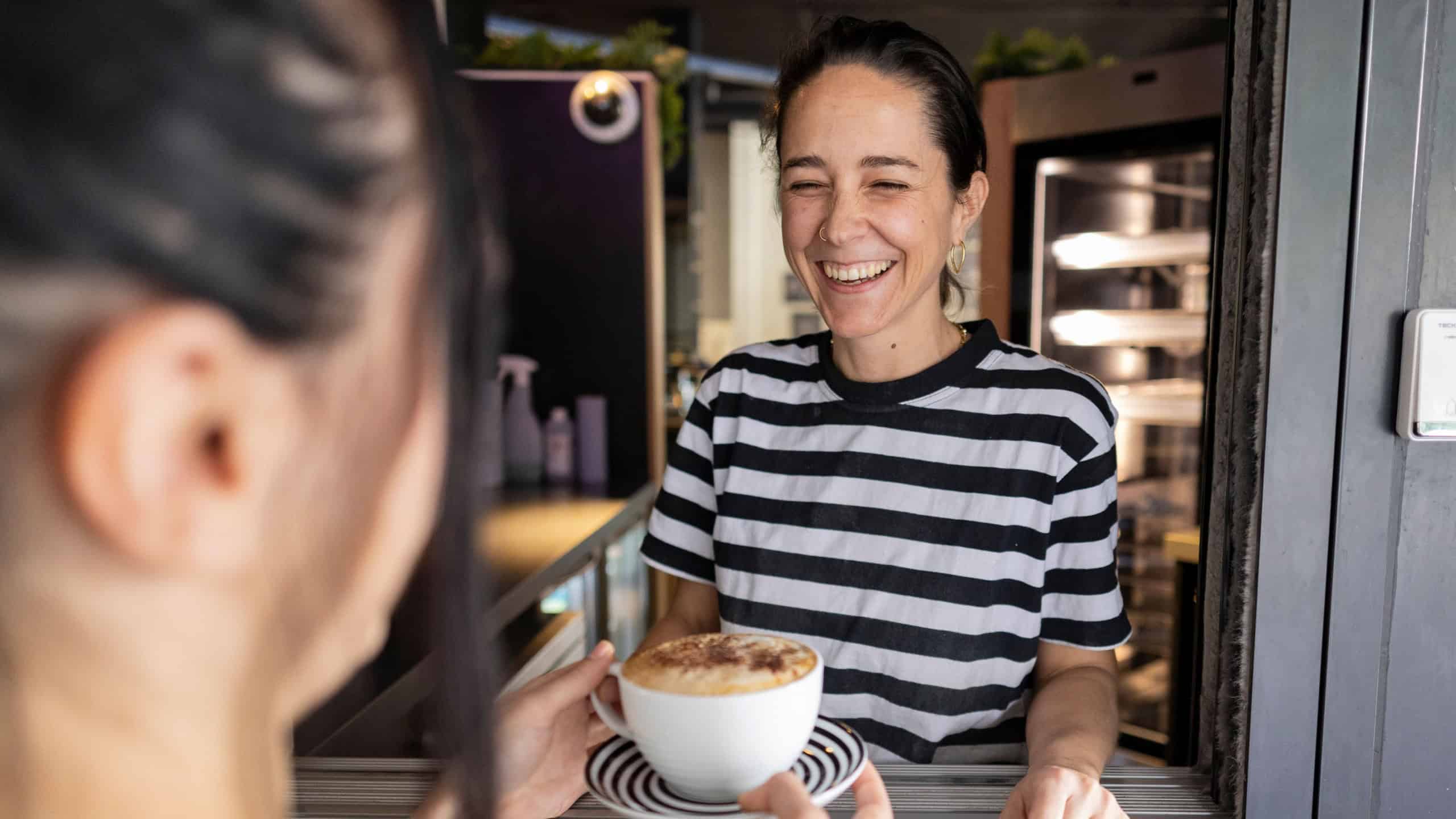 Happy waitress serving a steaming cup of cappuccino to a delighted customer, sharing a cheerful moment during a bustling workday in a trendy london cafe filled with vibrant energy
