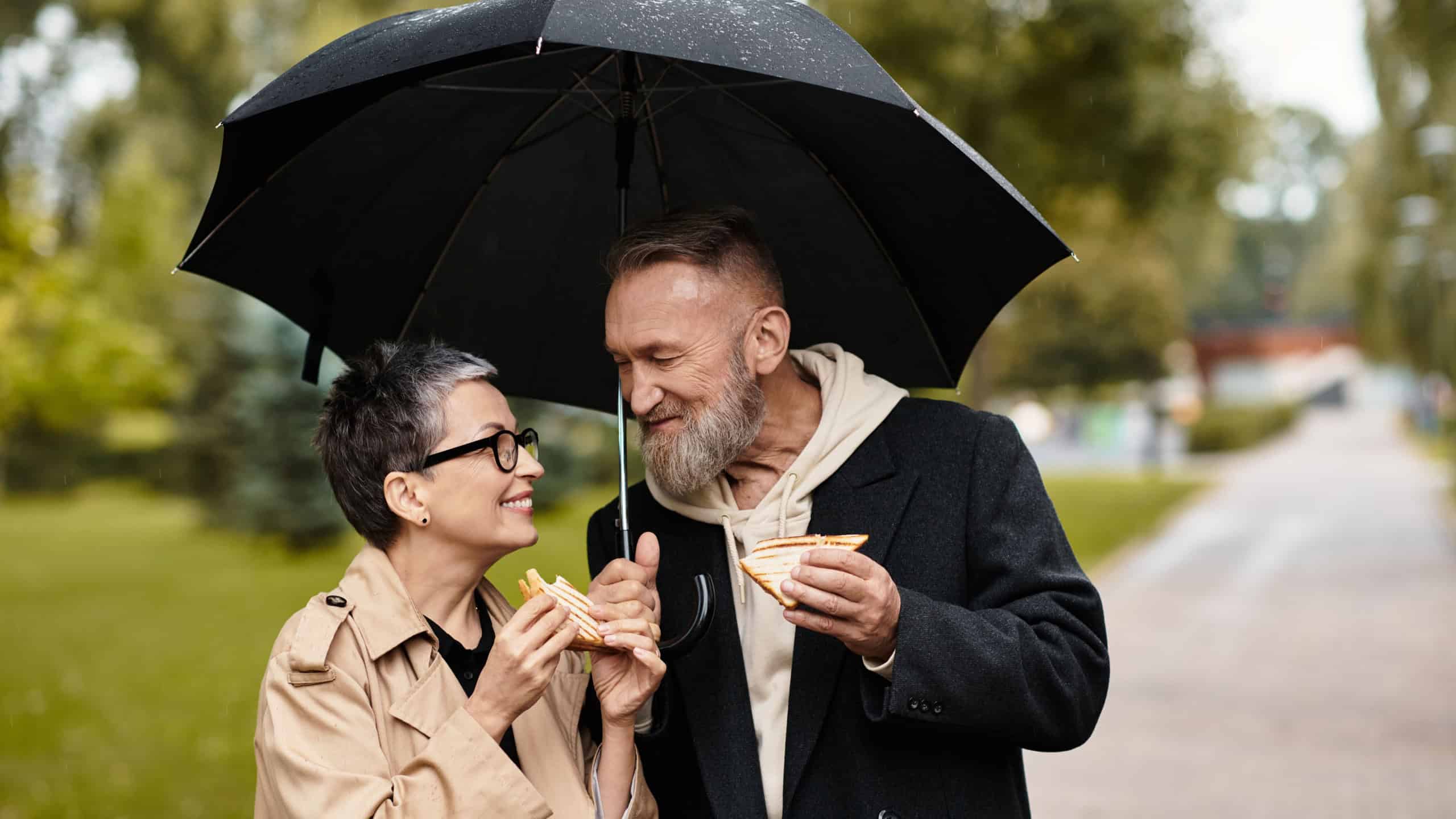 A loving couple enjoys sandwiches together while staying dry under a shared umbrella during a rain.