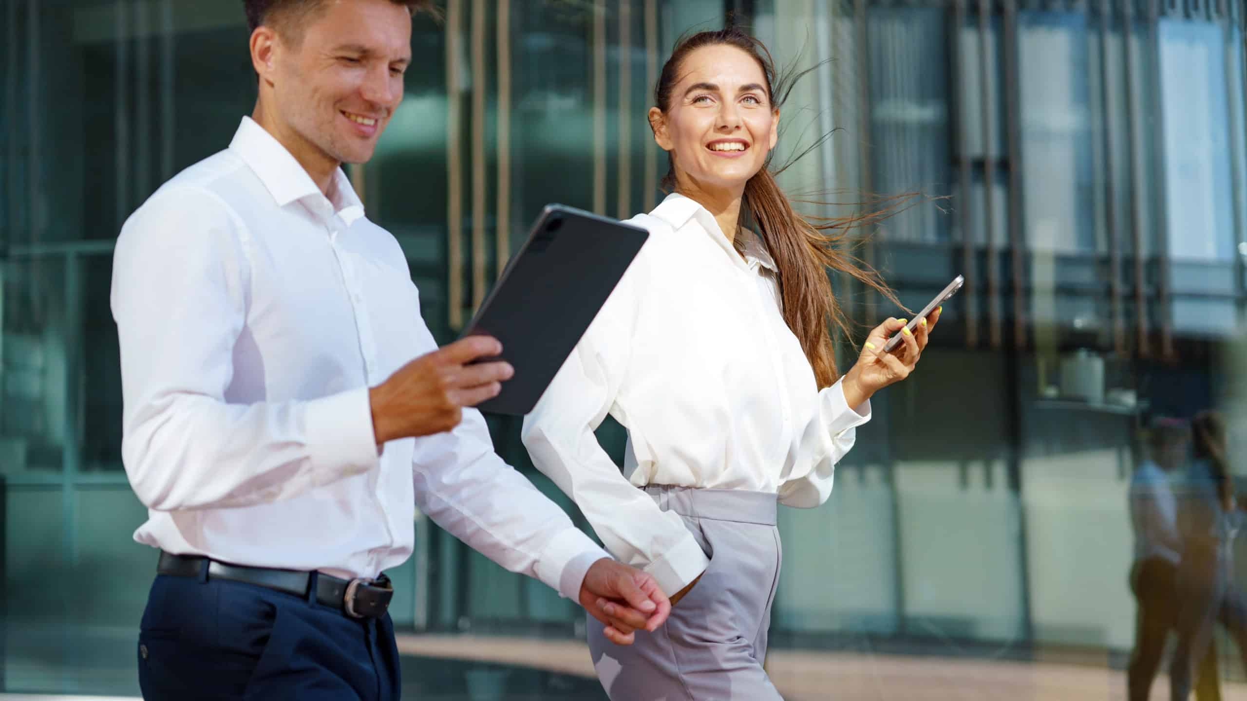Professionals in Modern Attire Engage With Technology While Walking Outside a Contemporary Office Building in the Afternoon Sun
