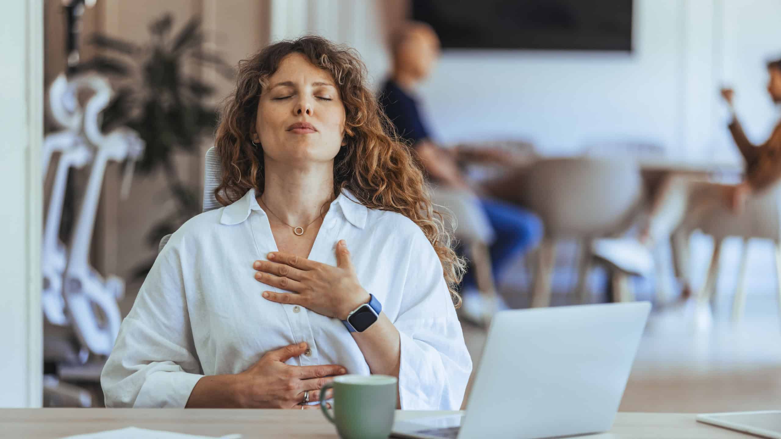 A professional woman seated at her desk practices mindfulness and relaxation, promoting workplace wellness. The scene emphasizes the importance of stress management and mental health in a busy office