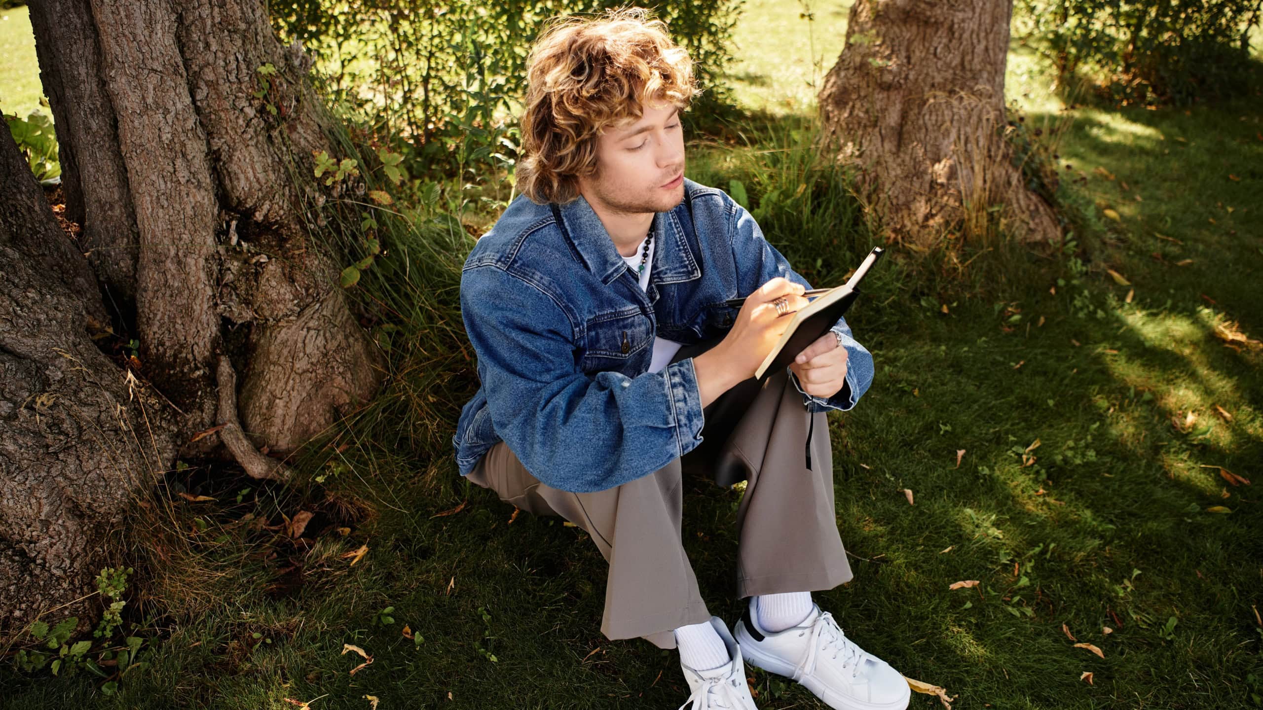 A young, charming man sits on grass, sketching in a notebook, surrounded by trees and nature.