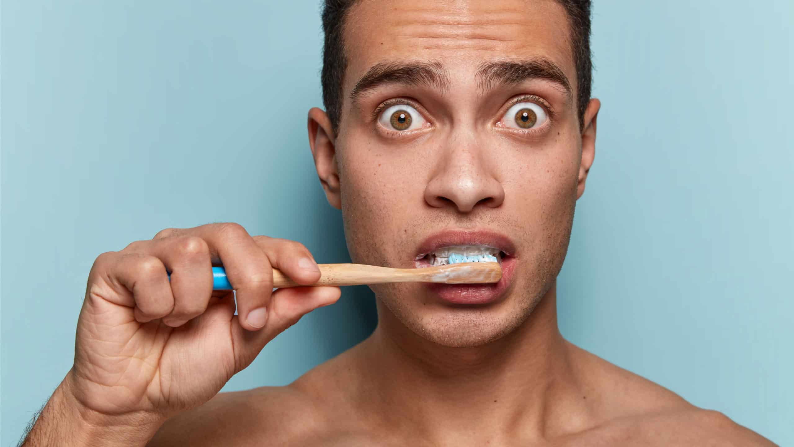 Teeth health concept. Stupefied young man with European appearance brushes teeth with bamboo toothbrush and toothpast, surprised to have caries or tooth decay, isolated on blue studio background
