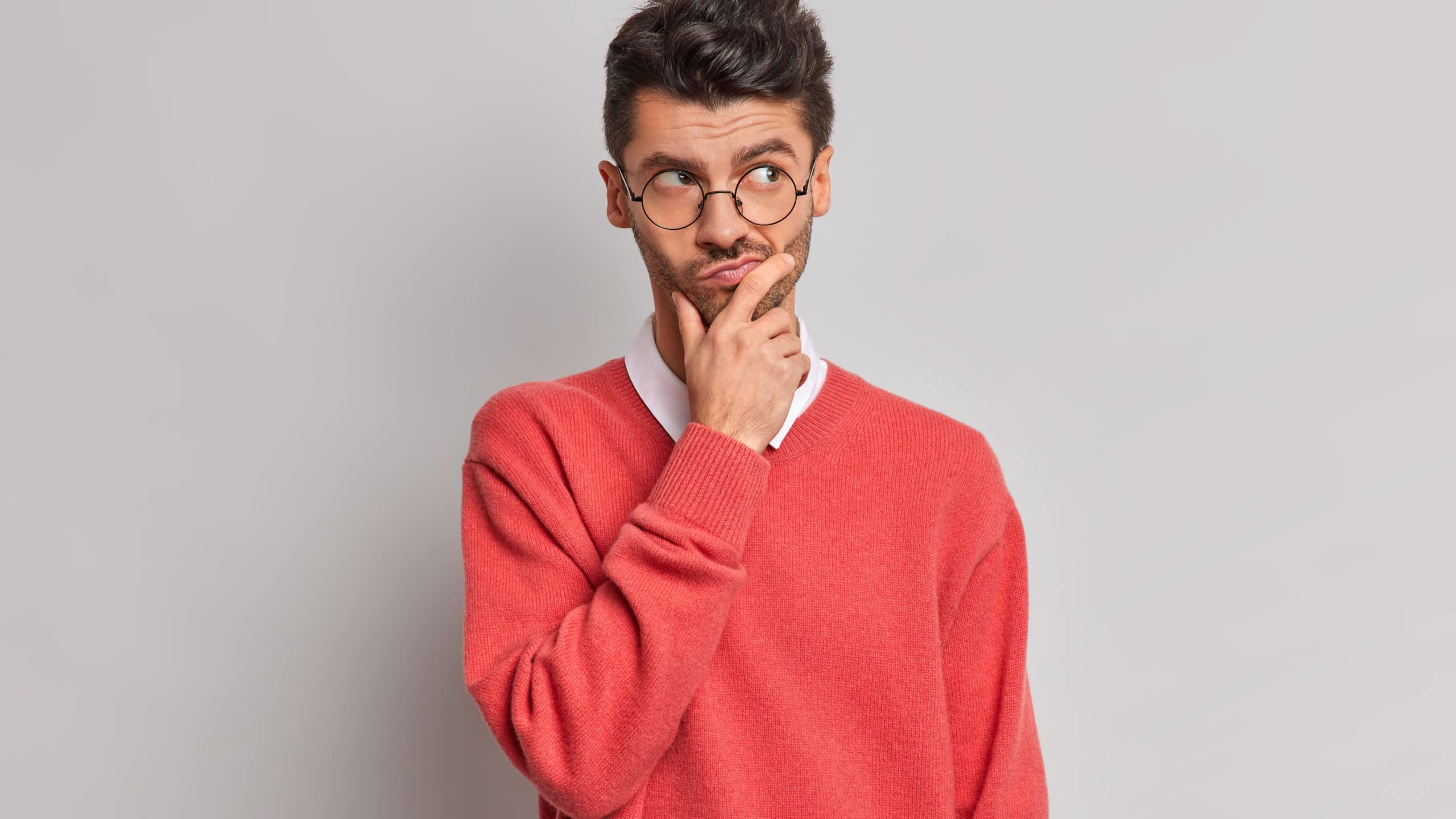 Photo of thoughtful handsome adult European man holds chin and looks pensively away tries to solve problem wears round glasses and red jumper isolated over grey background. Let me think about it