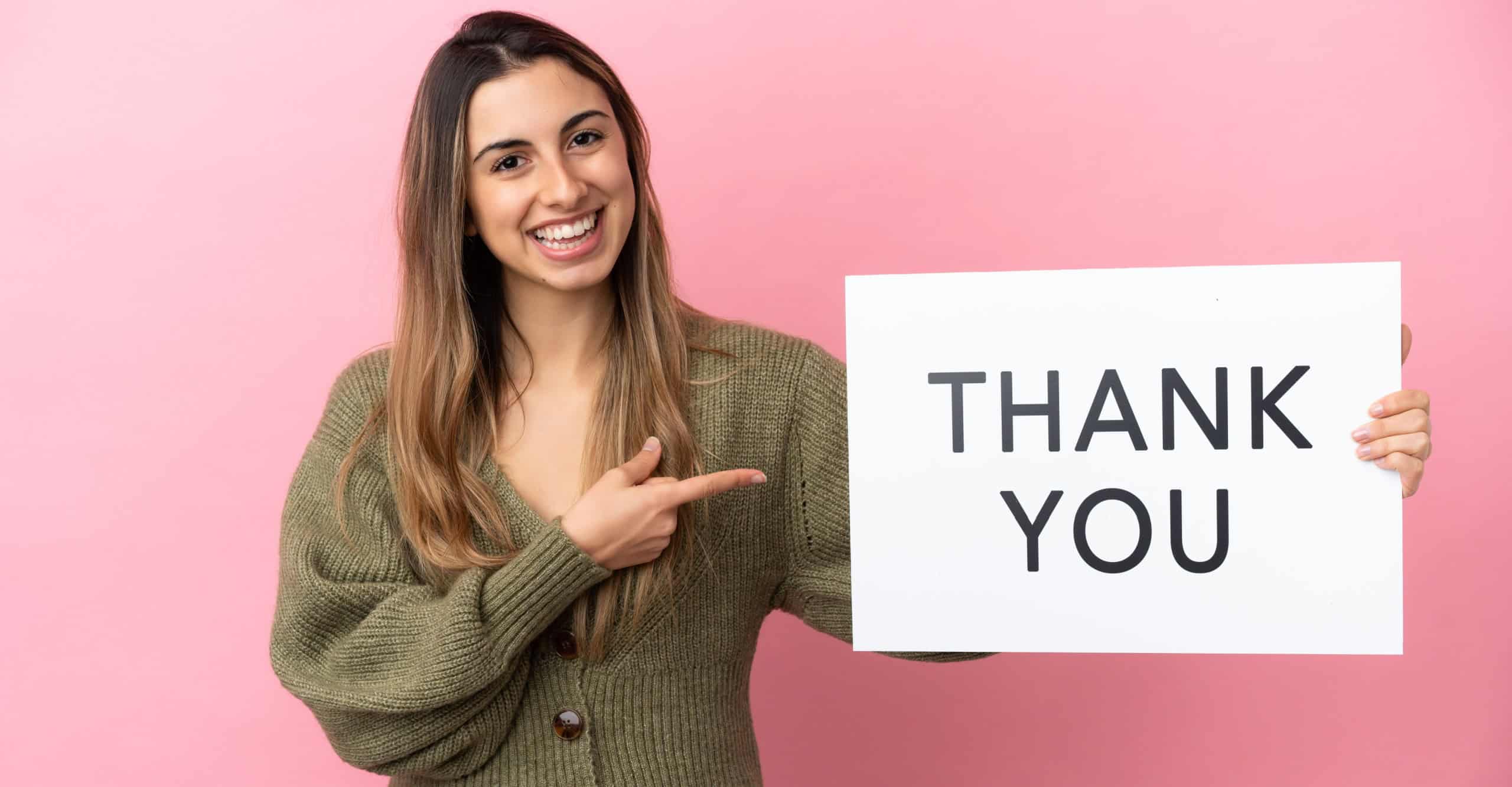 Young caucasian woman isolated on pink background holding a placard with text THANK YOU and pointing it