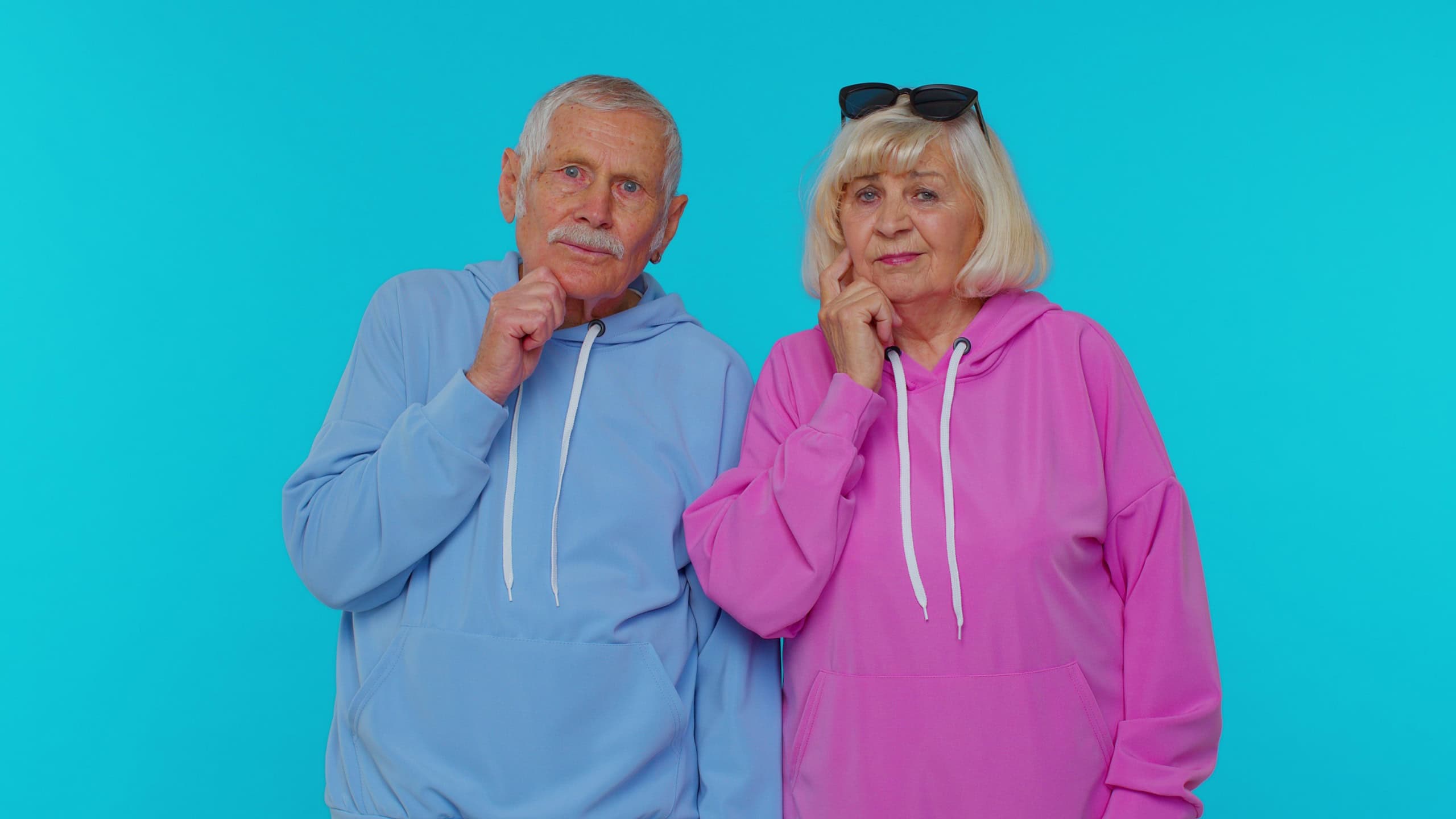 Thoughtful clever senior couple man woman grandparents rubbing chin, looking aside with pensive expression, pondering a solution, doubting question. Elderly grandfather grandmother on blue background
