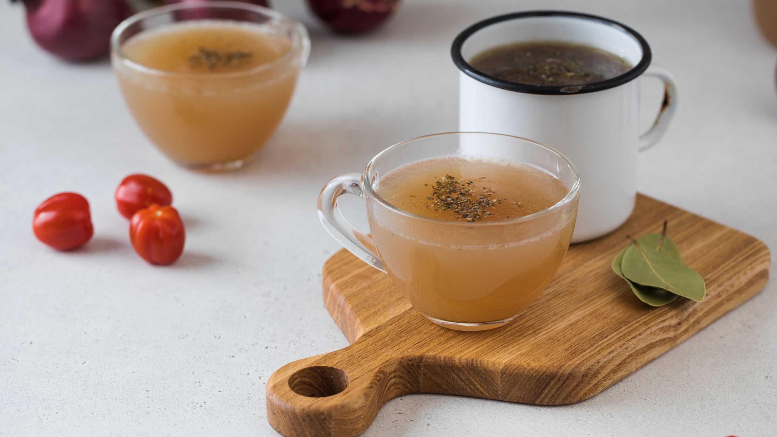 Homemade bone broth in mugs with spices on a wooden stick with bay leaf, tomatoes and onions.