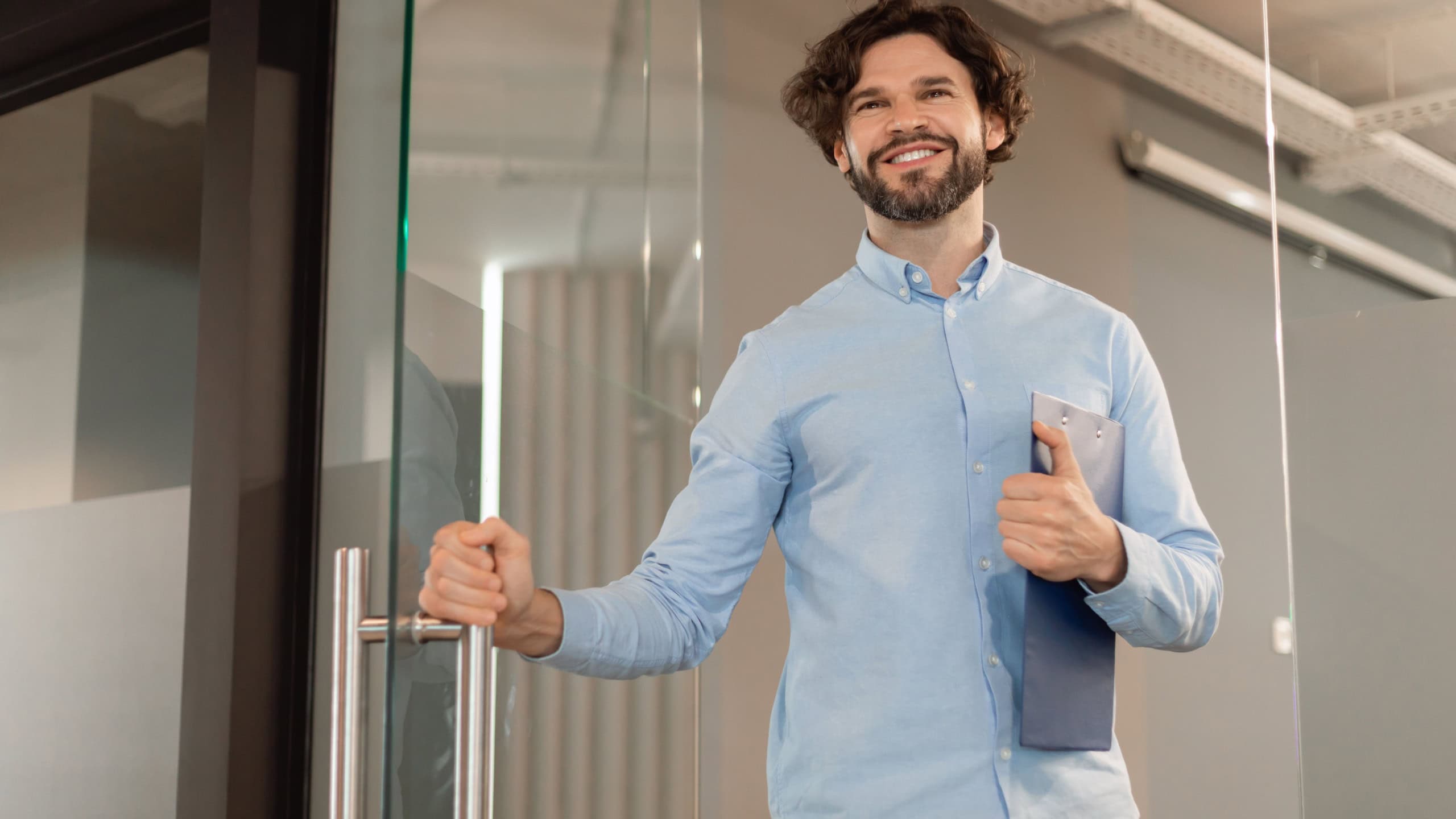 Portrait of confident smiling middle aged business man in shirt opening glass door walking in modern office and smiling, holding clipboard and handle, free copy space, low angle view