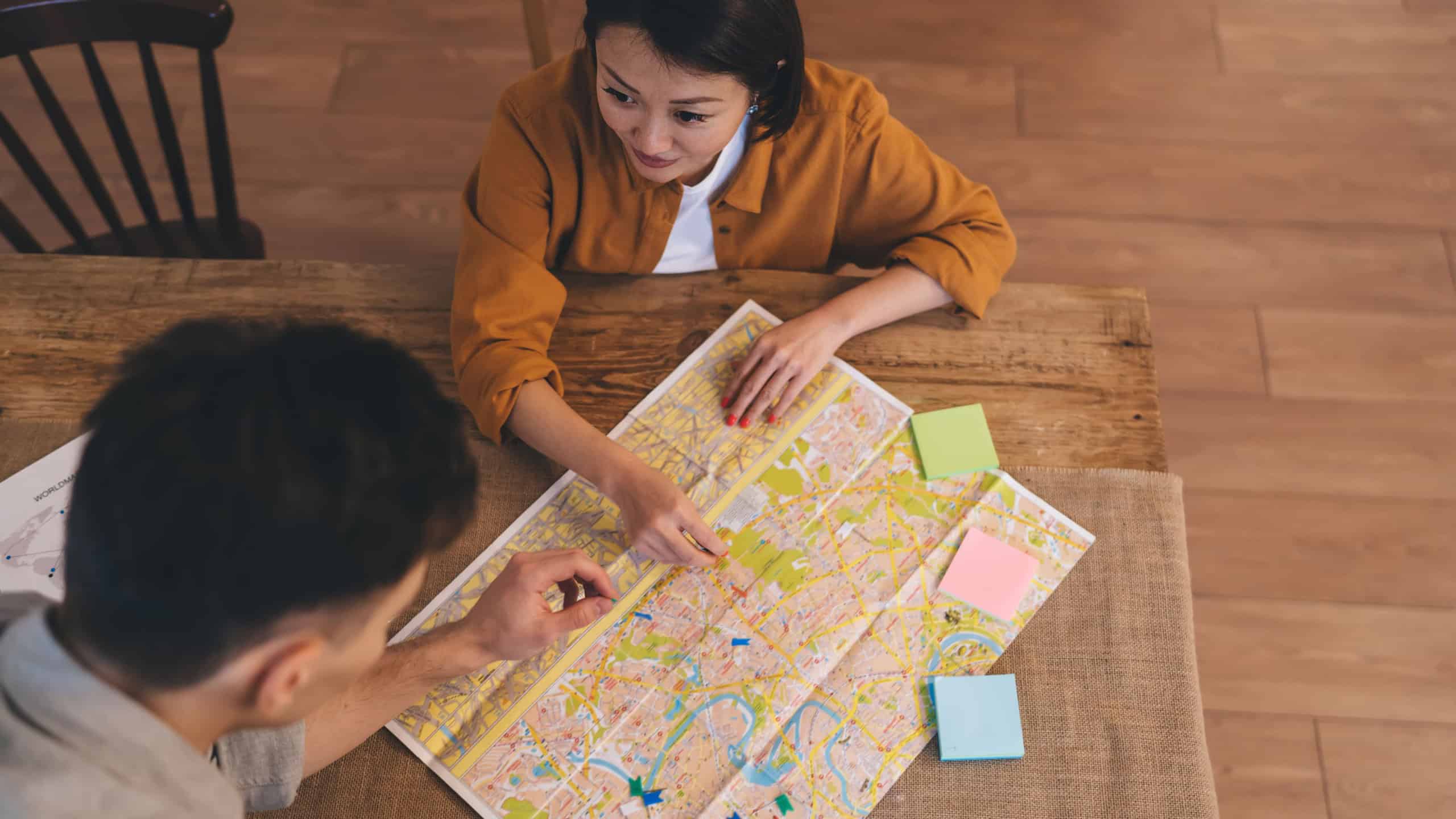Top view of couple choosing famous places on city map at table at home. Asian woman looking on boyfriend. Persons planning vacation or journey. Wooden desk with adhesive paper and notice board pins