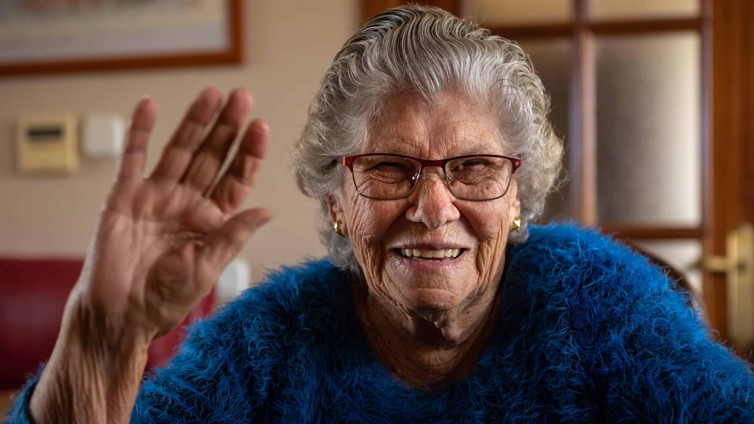Portrait of happy excited caucasian 90s woman enjoying video call talk, waving hand hello and looking at camera of webcam at living room. Happy senior retired lady or grandmother at home.