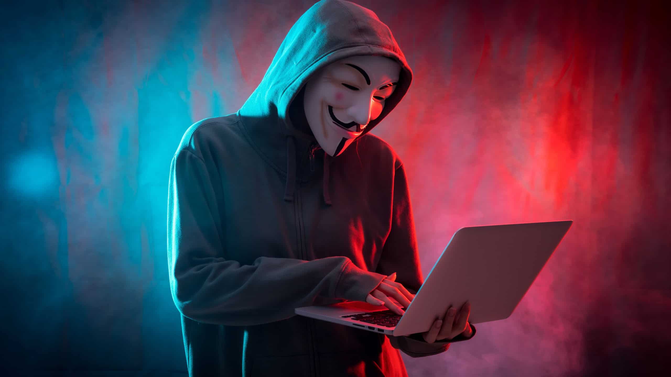 Hacker with anonymous mask with a computer and with a background of smoke and colored led