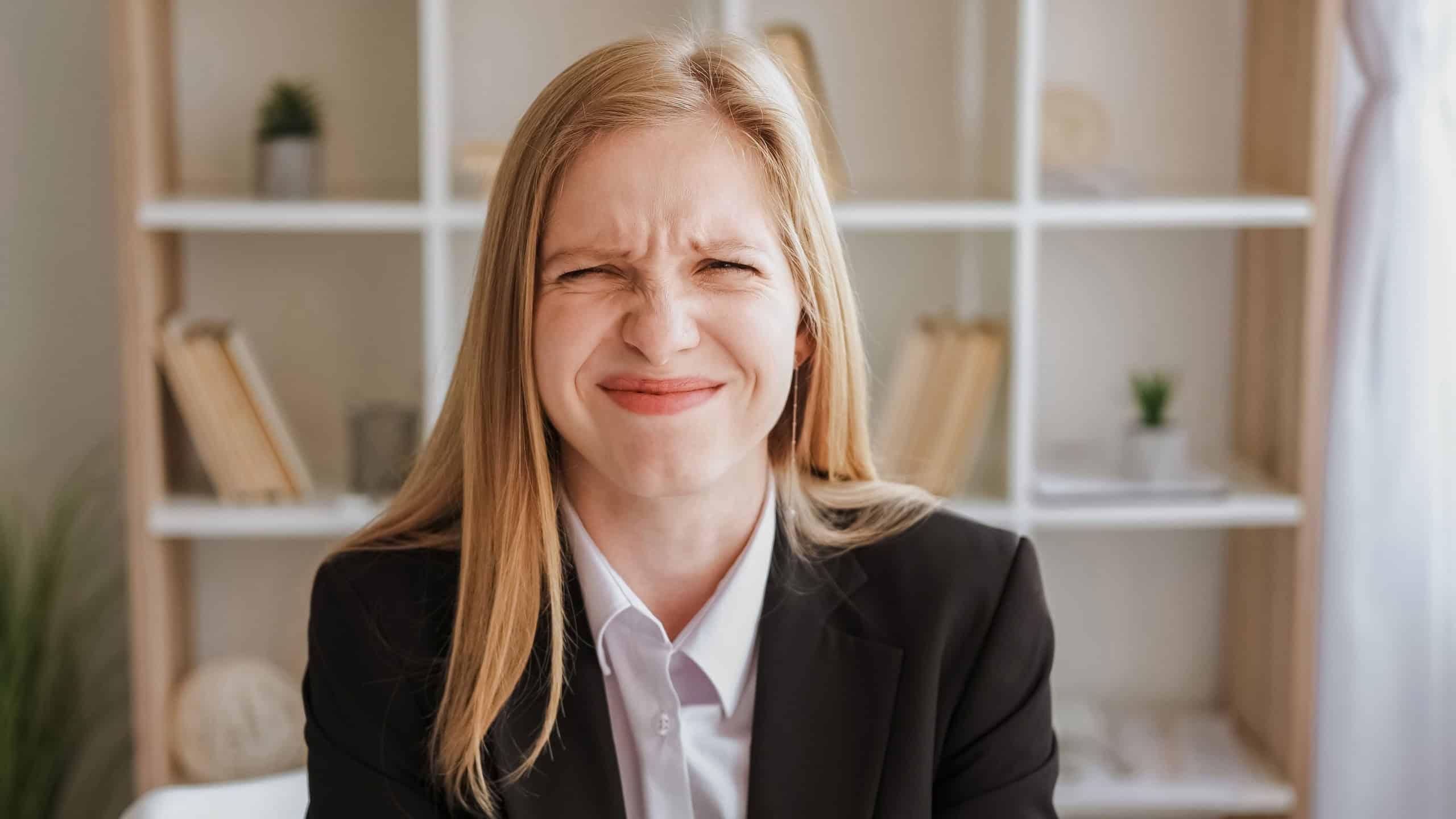 Embarrassed expression. Mistake fail. Funny confusion. Cute ashamed blond young woman in business suit with wincing grimace in light interior.
