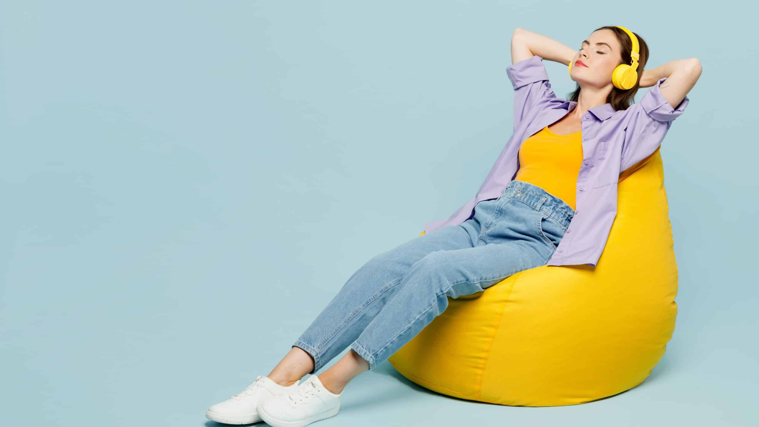 Full body side view young happy woman wears purple shirt yellow t-shirt casual clothes sit in bag chair listen to music in headphones isolated on plain pastel light blue background. Lifestyle concept
