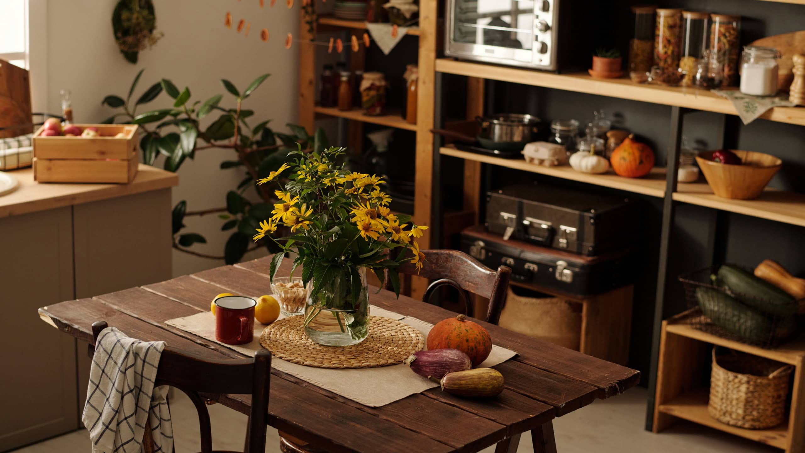 Wooden table with bunch of wildflowers in vase, vegetables, mug and two chairs standing in the center of spacious kitchen of country house