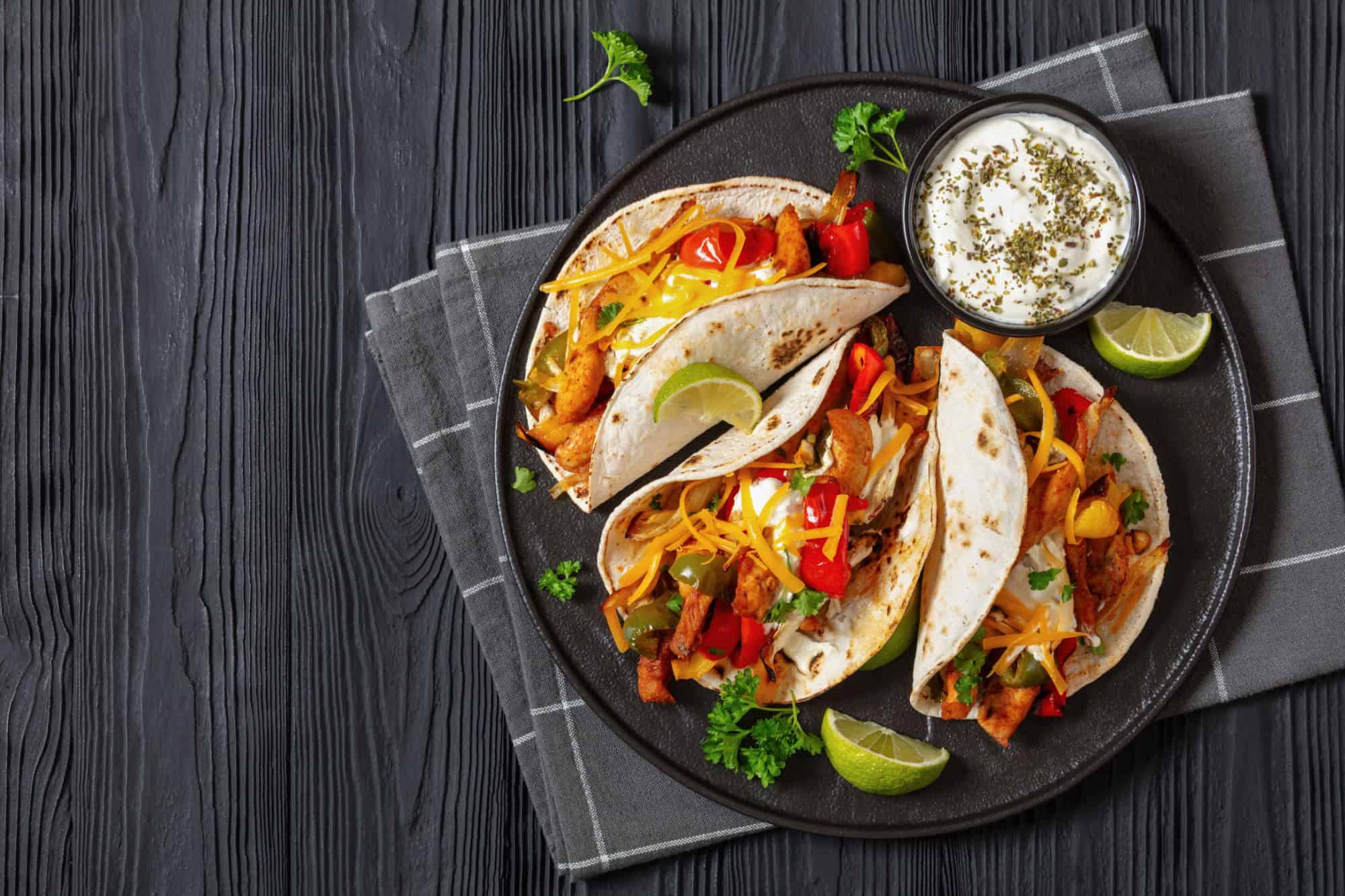 baked tex-mex chicken fajitas with mixed sweet pepper, onion, sour cream, shredded cheese and white corn tortillas on black plate on black wooden table, horizontal view, flat lay, free space