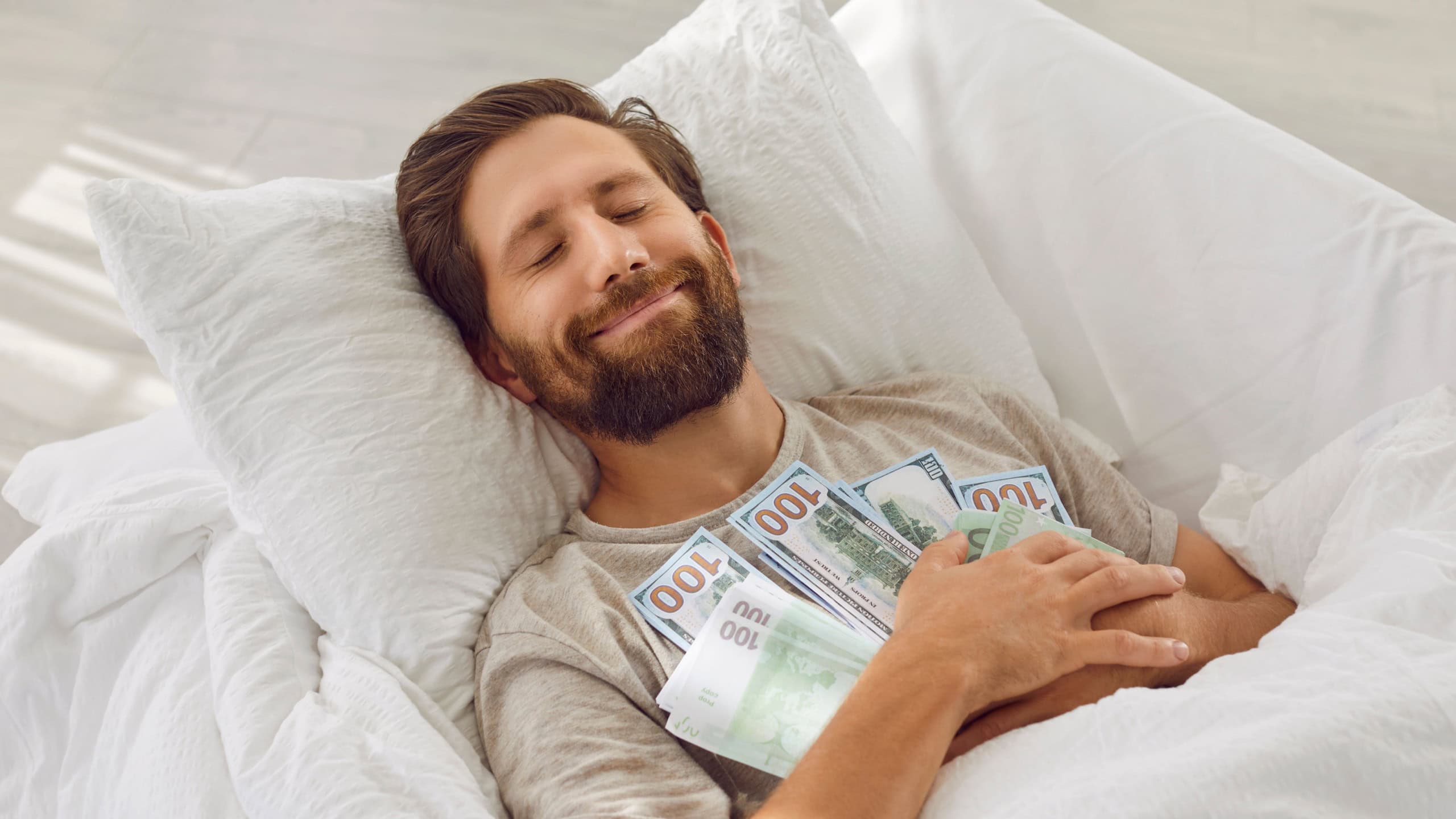 Young rich man sleeps with pile of cash and smiles with satisfaction in his sleep. Caucasian man sleeping in bed holding pile of cash dollars and euros keeping his savings at home. Wealth concept.