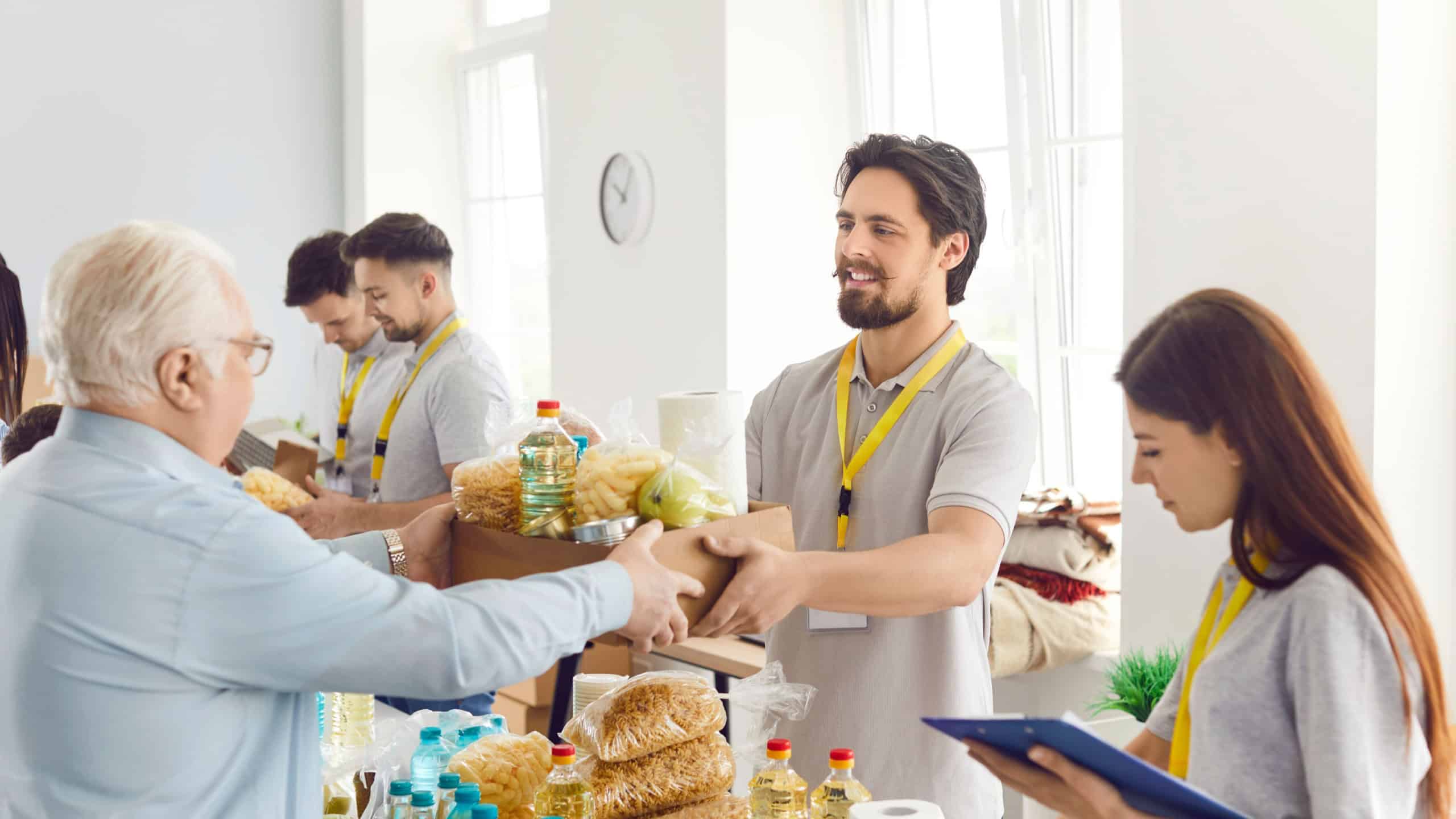 People getting food aid at a charity center. Helpful volunteers give a senior man a box with pasta, oil, canned food and other stuff. Donation, poverty, help concept