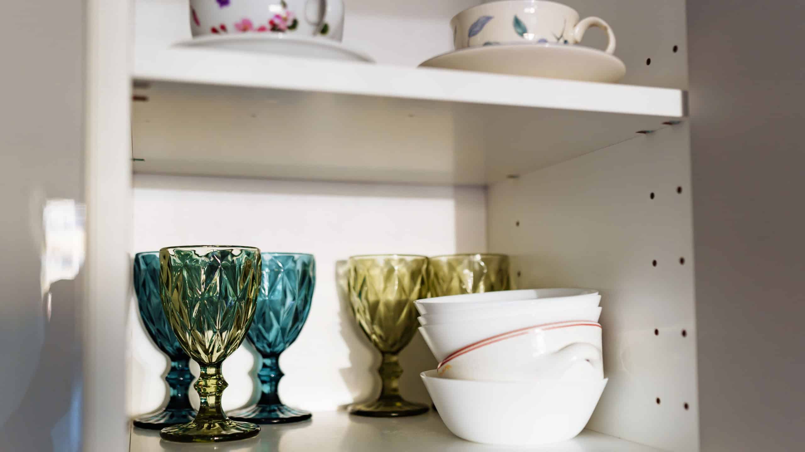 Clean dishes, colored glasses, cutlery on white shelf in kitchen cabinet.