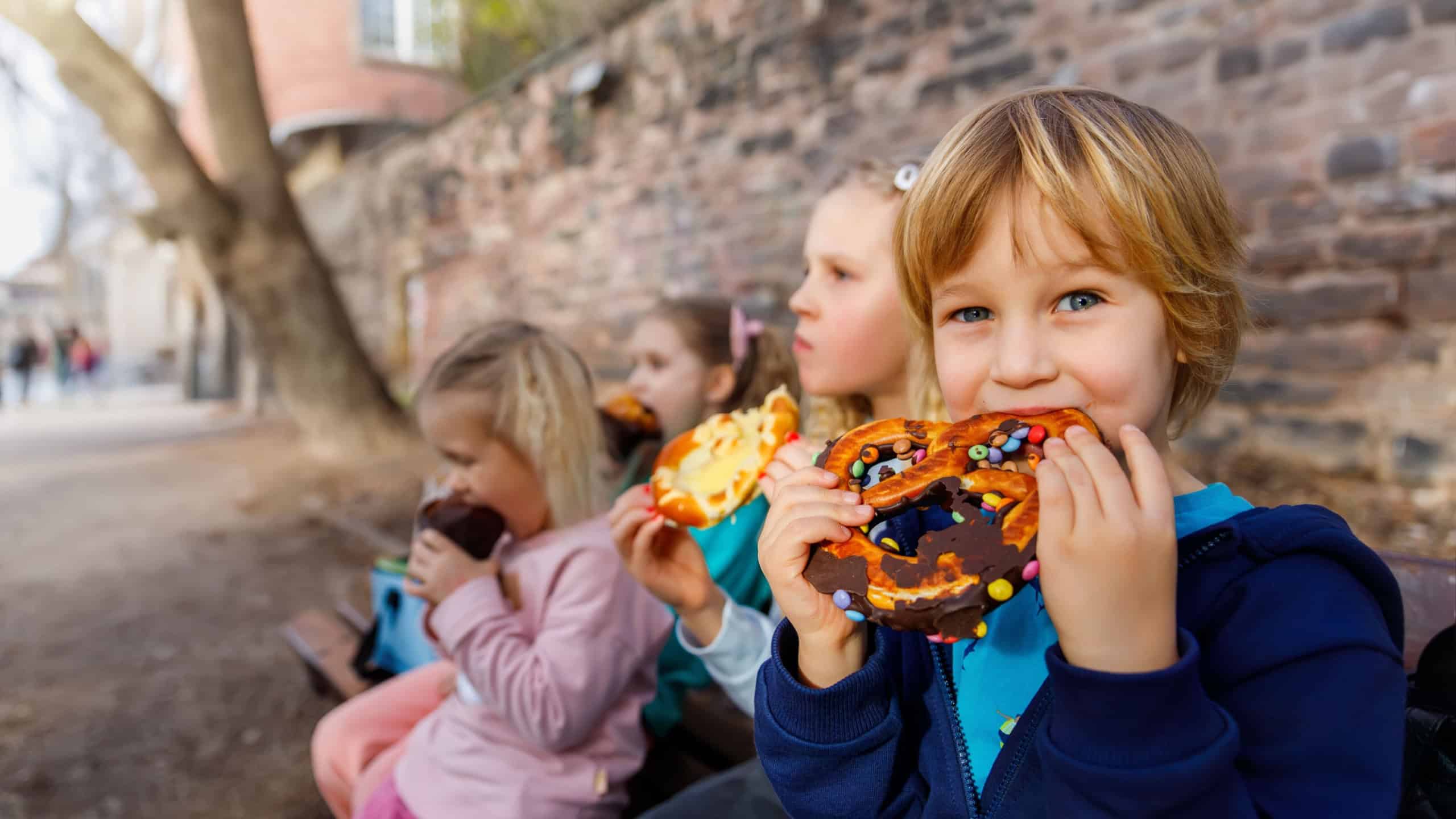Portrait group happy funny playful blond children enjoy eating pretzels chocolate toppings at city street outdoor park. Scenic fun moment capturing kids childhood joy snacking on colorful treats