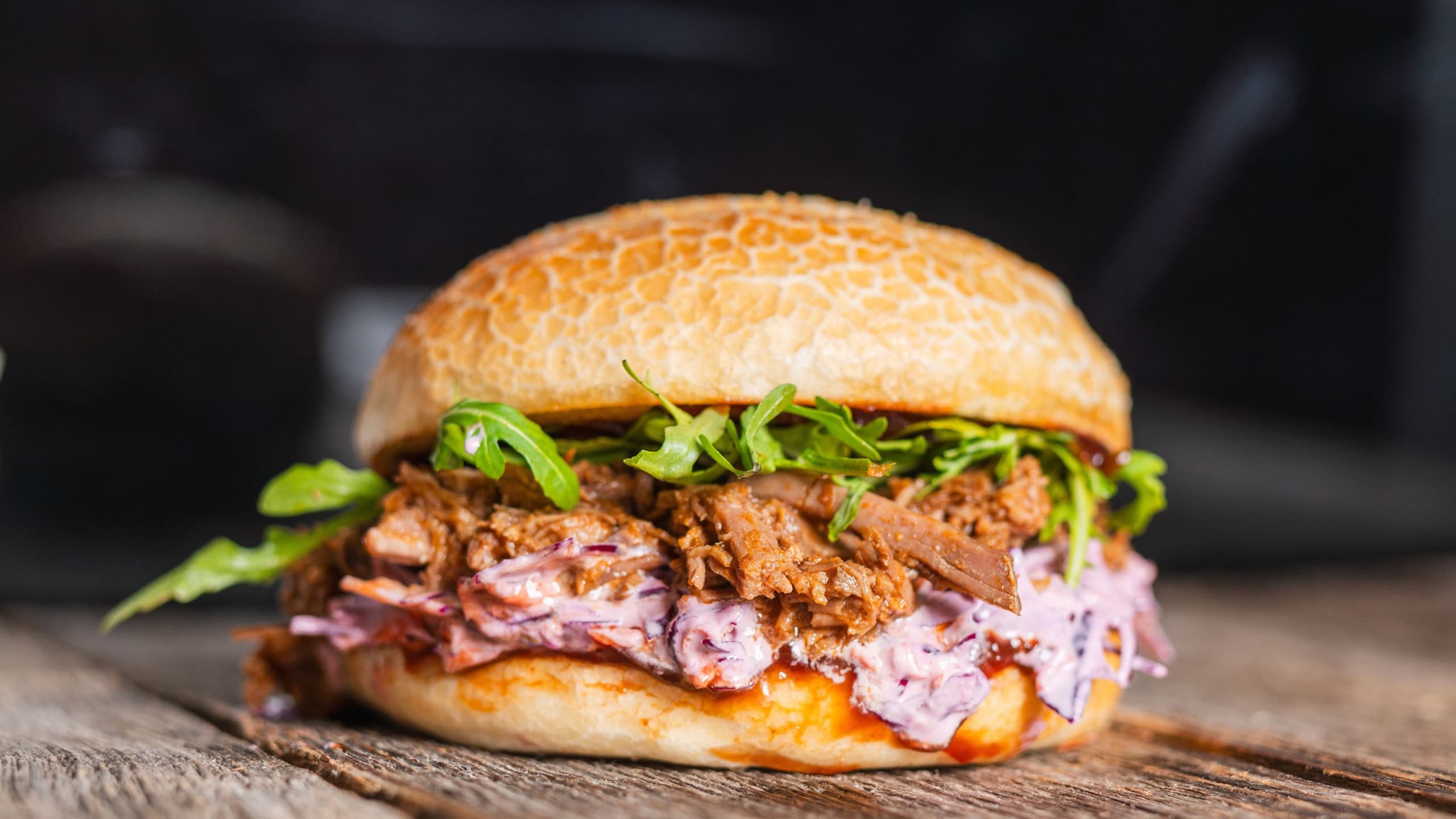 Pulled beef burger with fresh arugula, coleslaw, and barbecue sauce, served on a rustic wooden table.
