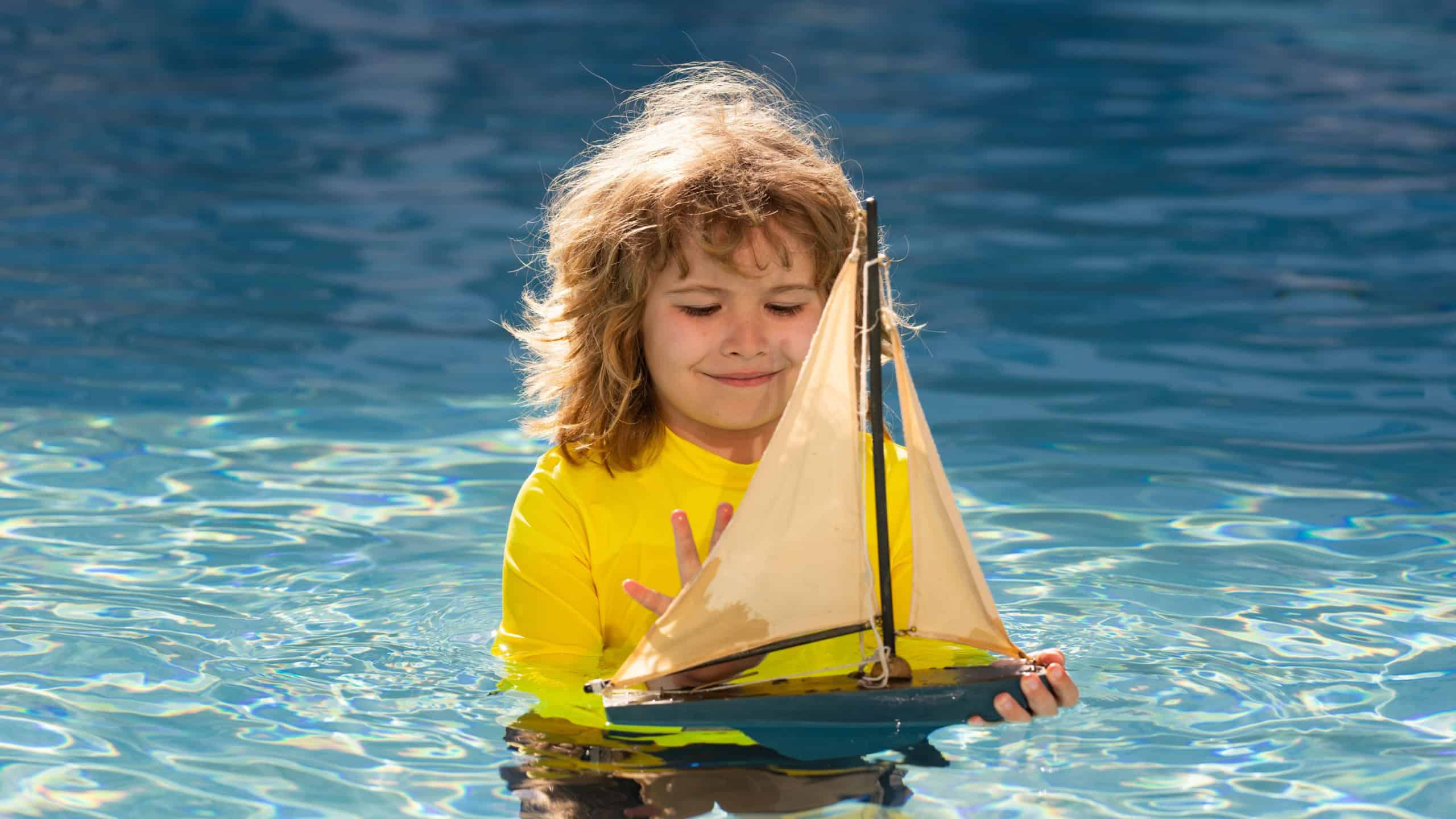 Kids travel concept. Kid playing with toy sailing boat on summer sea background. Travel and summer vacations. Child playing with toy boat in water on the beach. Outdoor summer activities for kids.