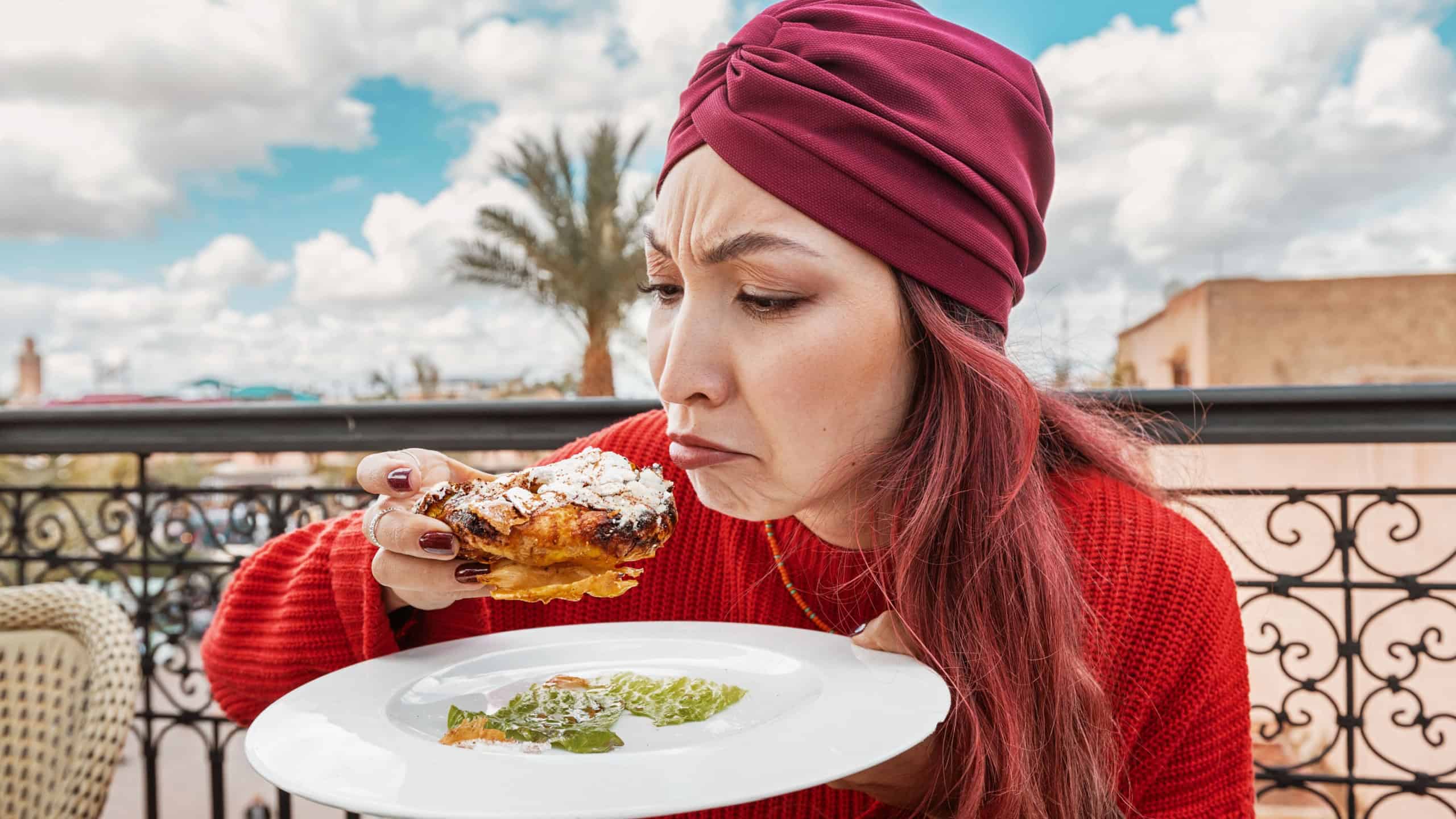 A woman on the terrace of a Moroccan restaurant tastes pastilla while looking at it with a suspicious and dissatisfied expression