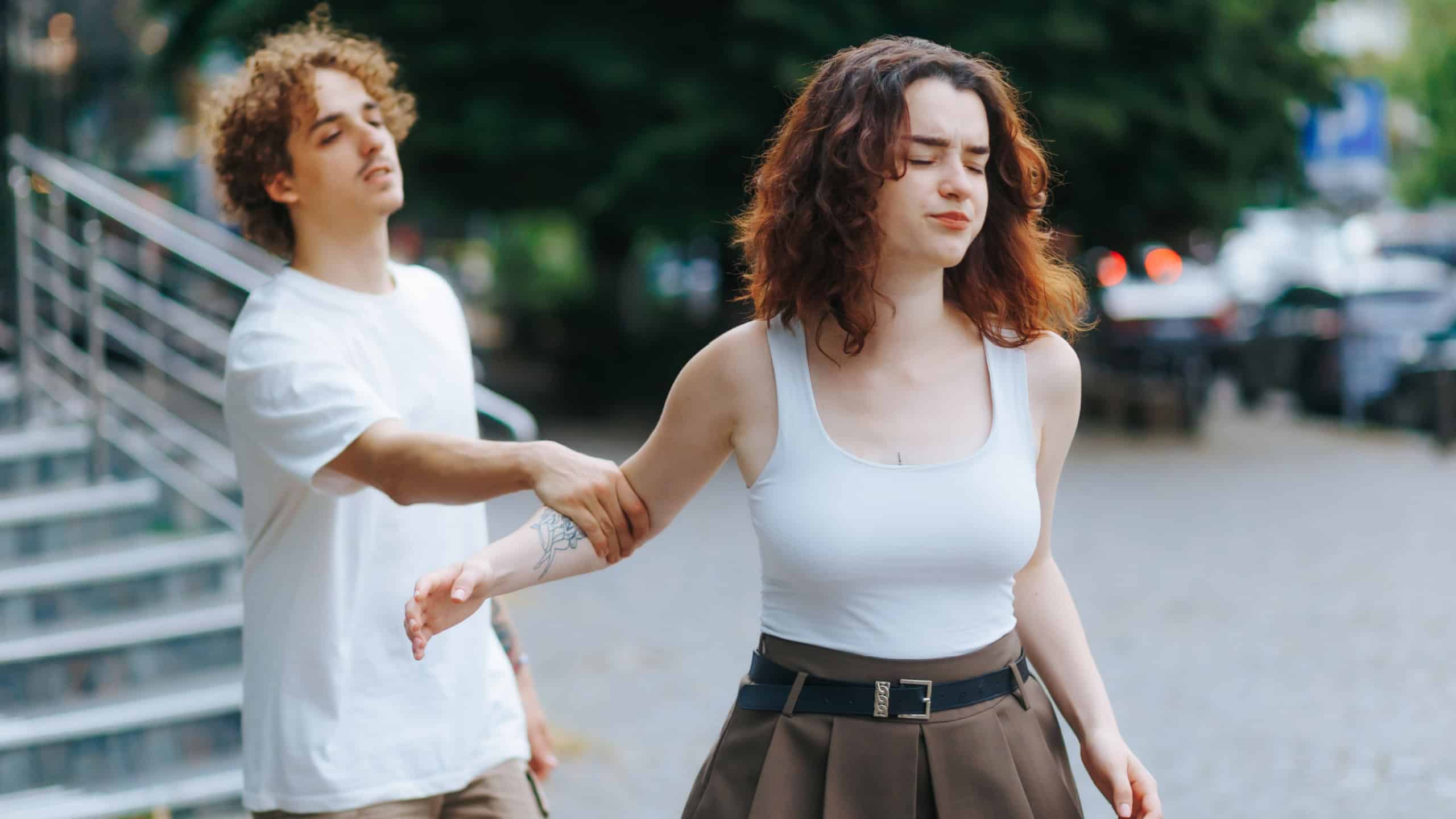 An emotional young man tries to talk to his girlfriend as she walks away angrily during a disagreement. Outdoor couple tension captured in a real urban moment.