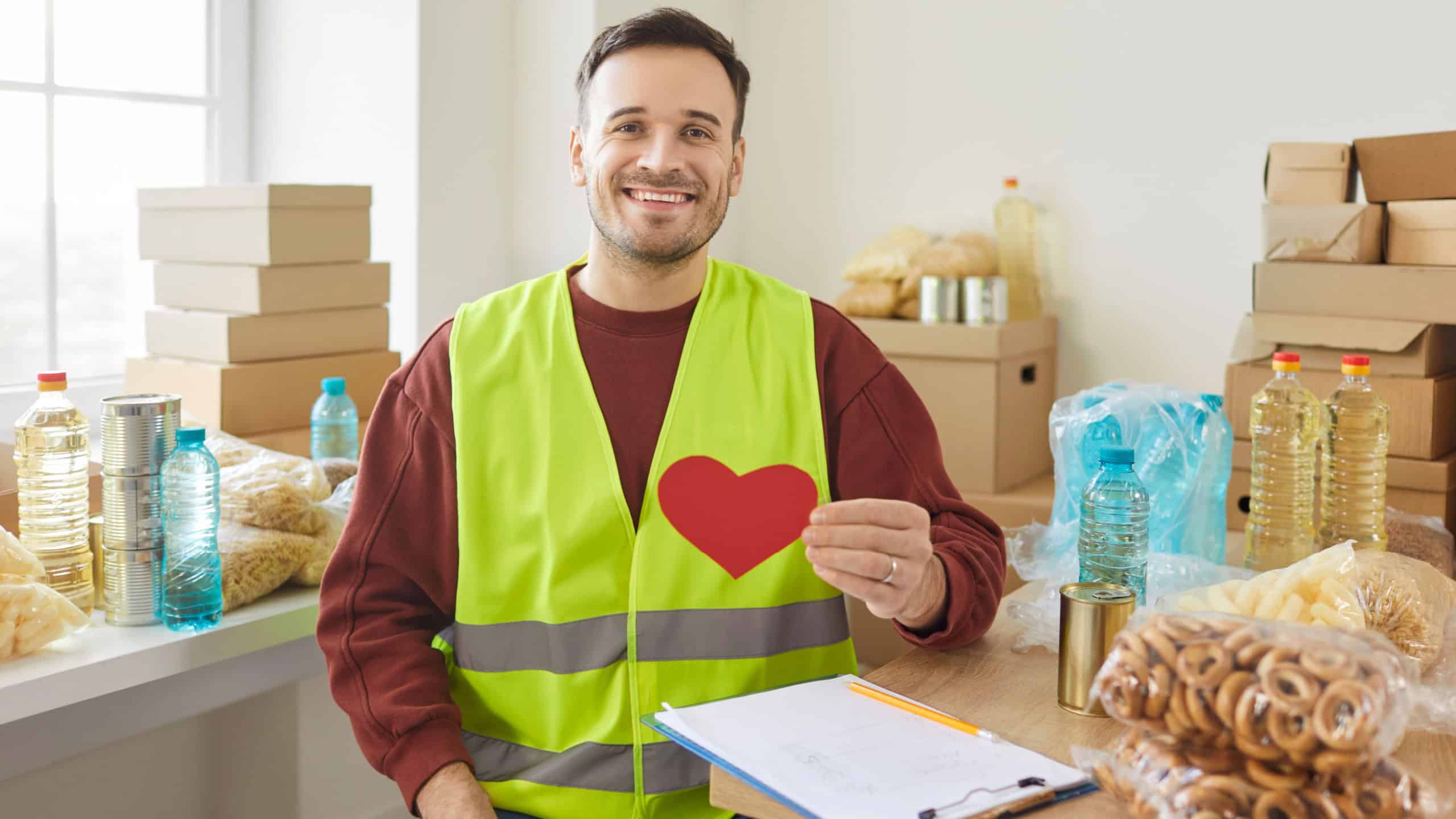 Portrait of smiling young man volunteer holding red heart in hands and looking at camera working at charity center. Male person volunteering in charitable foundation. Humanitarian aid concept.