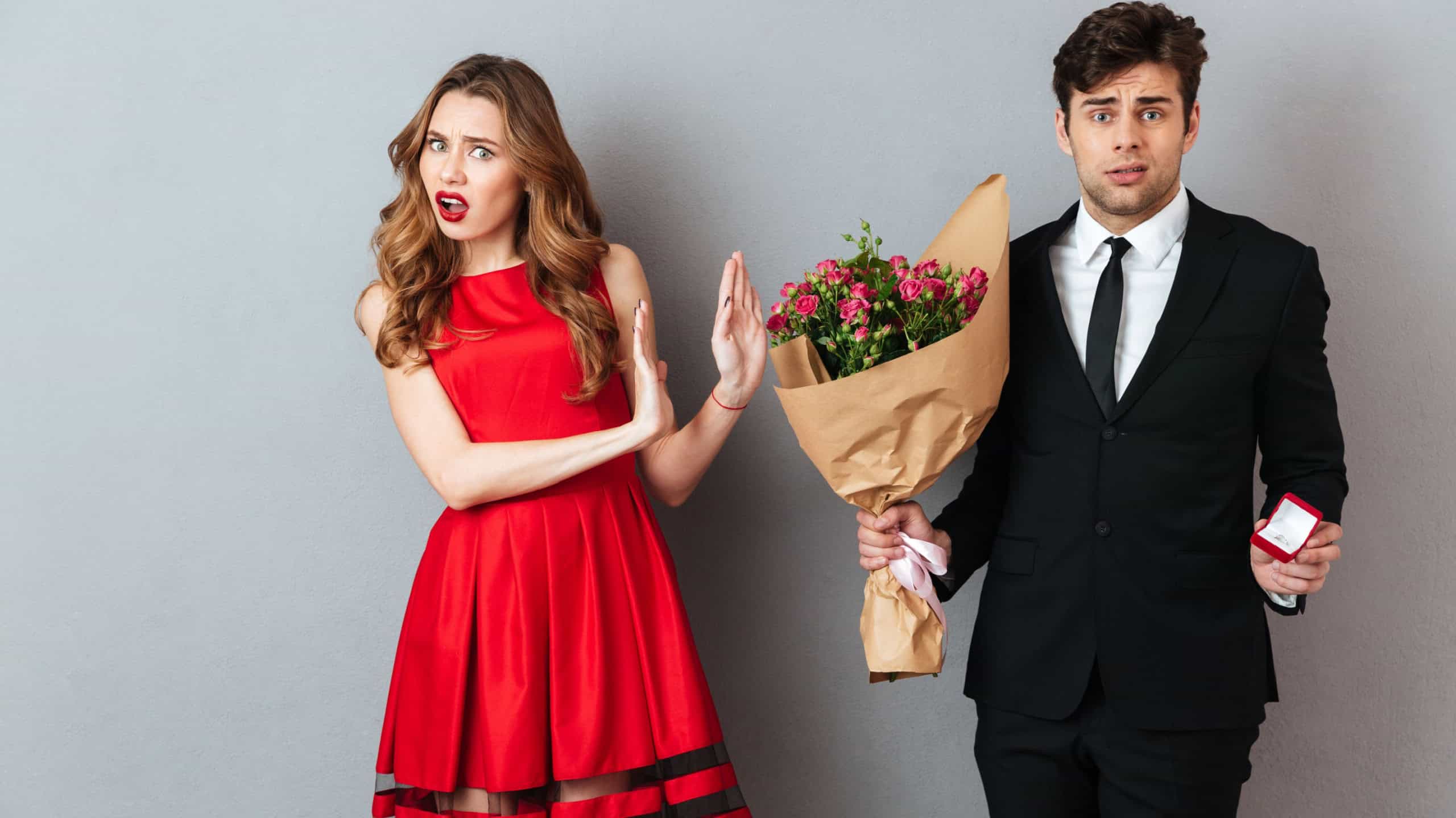 Portrait of a frustrated man proposing to a girl with flowers and an engagement ring and getting denied over gray wall background
