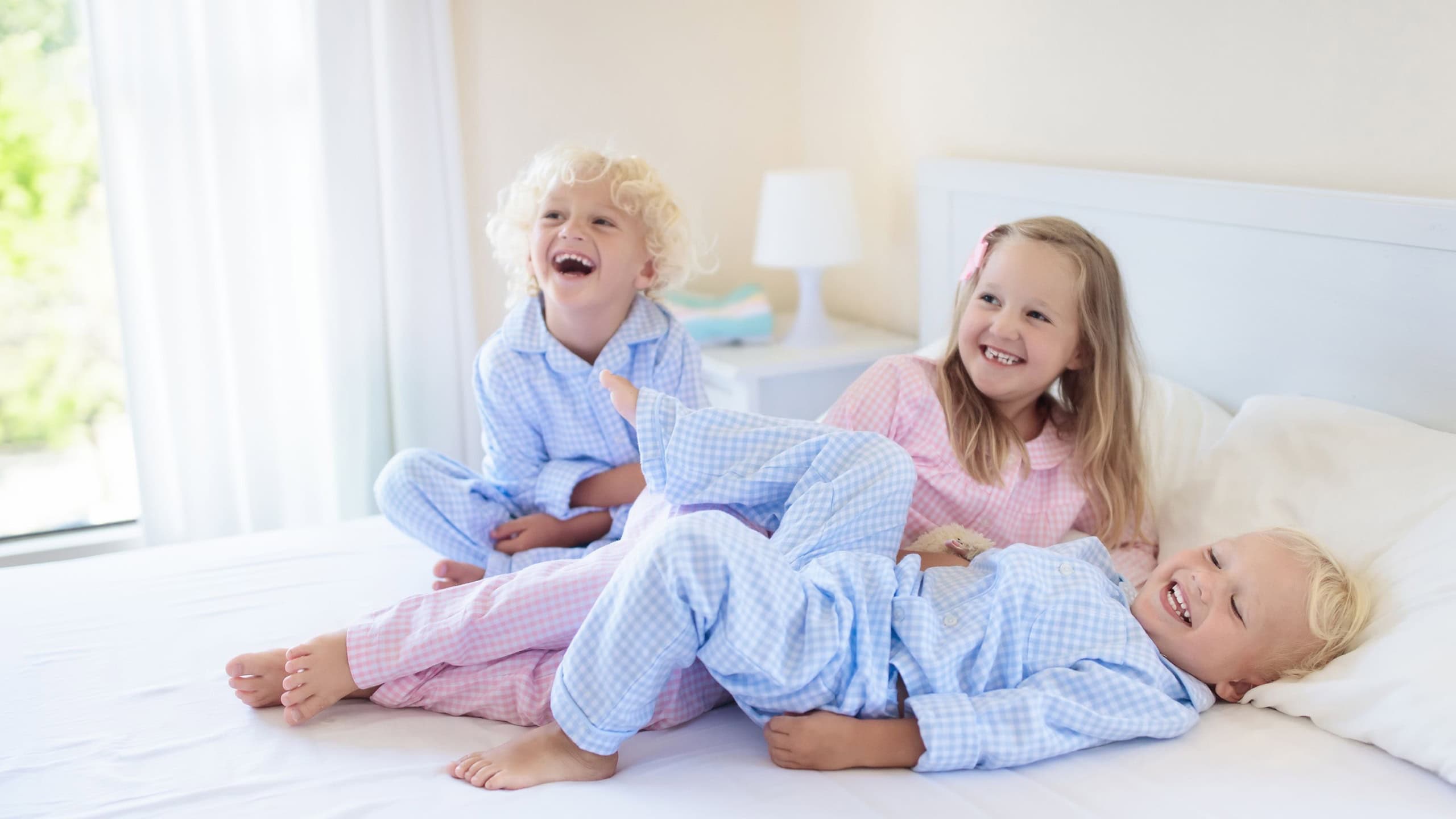 Kids playing in parents bed. Children wake up in sunny white bedroom. Boy and girl play in matching pajamas. Sleepwear and bedding for child and baby. Nursery interior for toddler kid. Family morning.