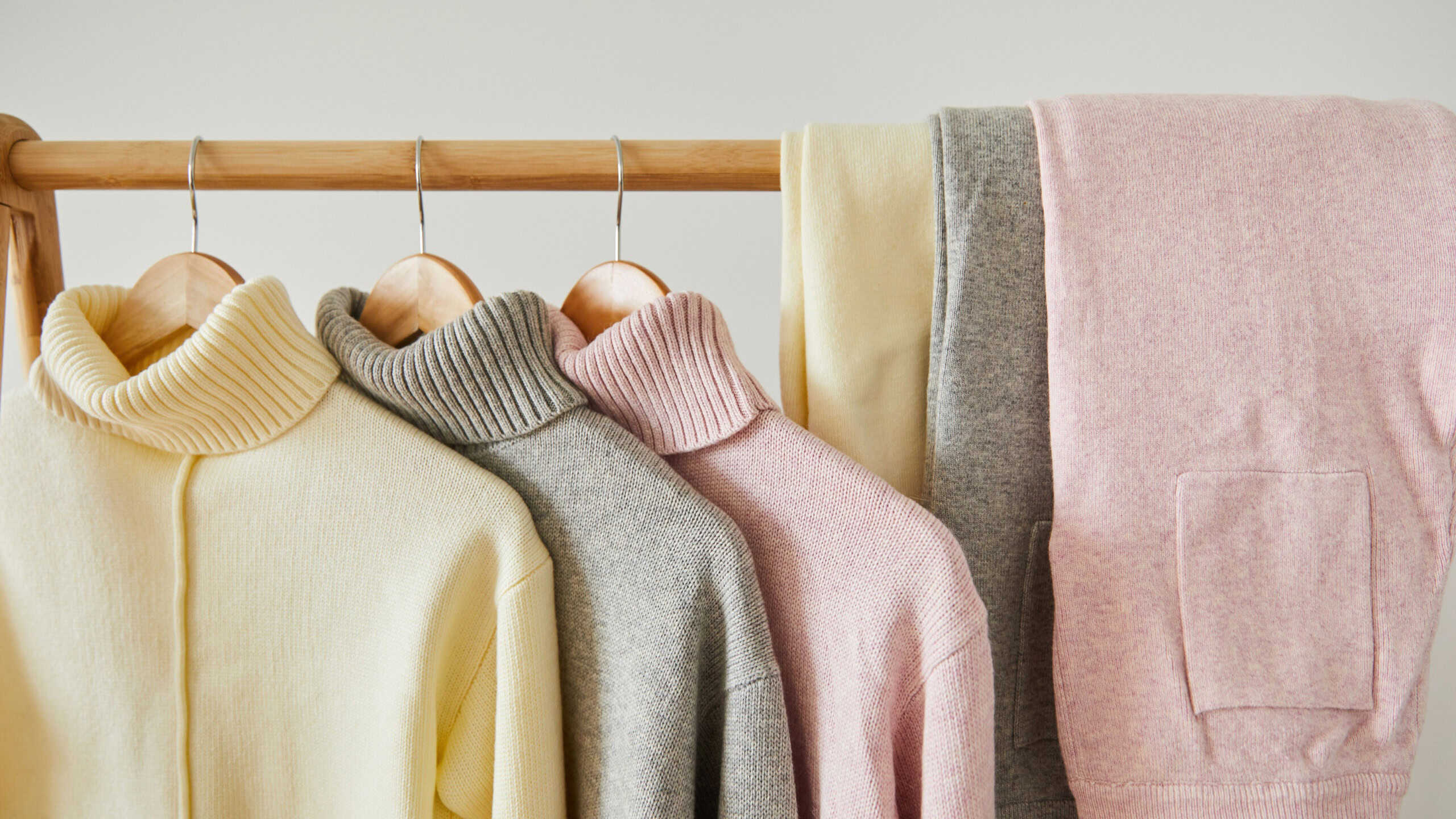 close up view of pink, beige and grey knitted soft sweaters and pants hanging on wooden rack isolated on white