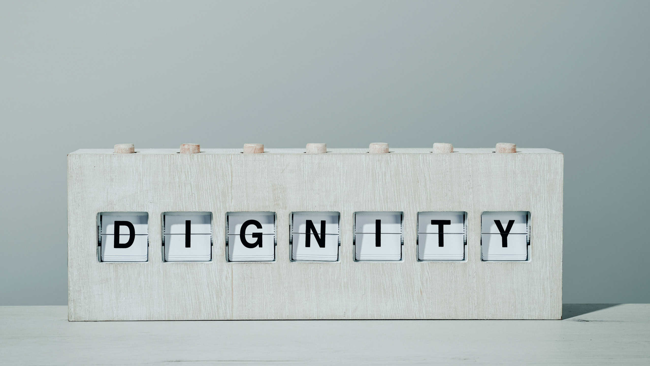 the word dignity in a rustic wooden split flap board, on a wooden surface in front of a pale gray wall