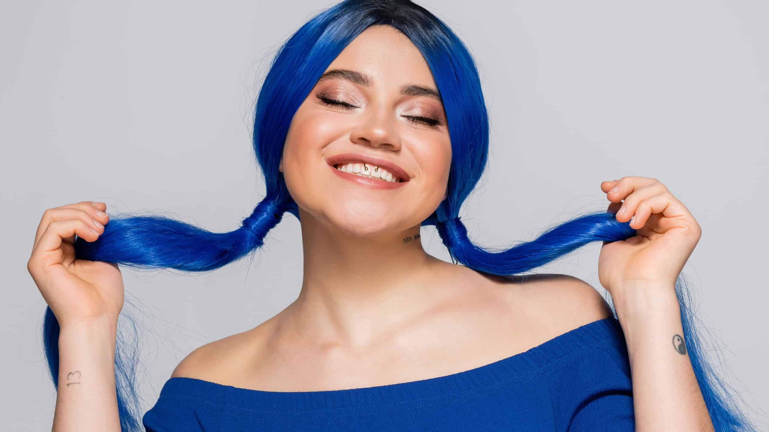 positivity and youth, tattooed woman with closed eyes and dyed hair smiling on grey background, hairstyle, blue hair, modern beauty, self expression, individualism