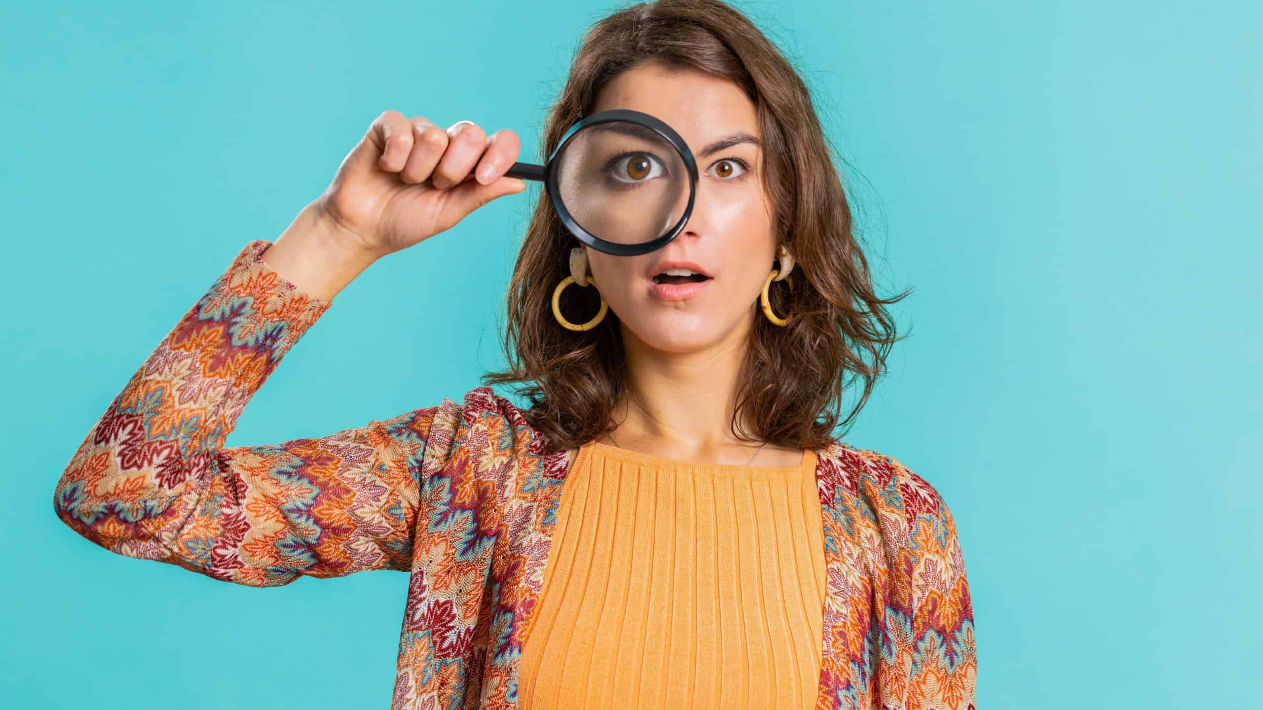 Investigator researcher scientist woman holding magnifying glass near face, looking into camera with big zoomed funny eyes, searching, analysing. Pretty young girl isolated on blue studio background