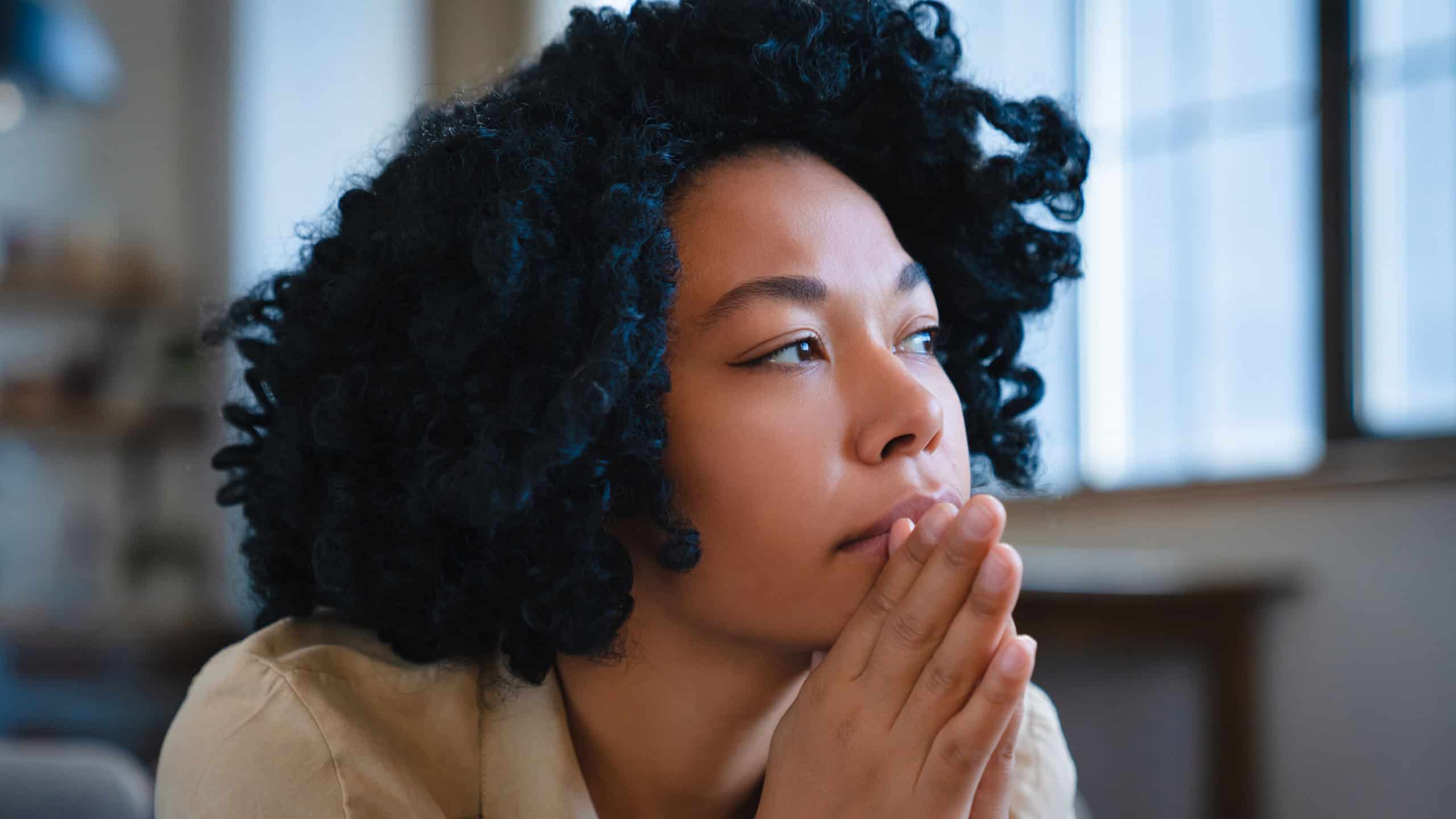 Discouraged pensive African American woman looking into distance thinking about ways to solve financial problems. Frightened black girl sits on sofa in living room having lost mood due to inflation