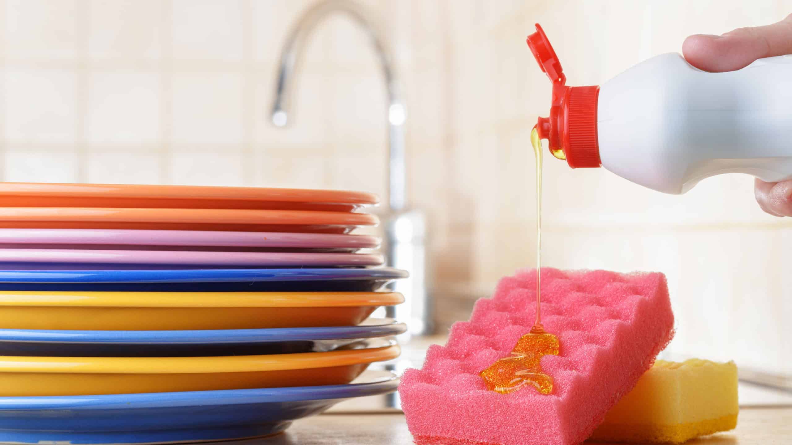 View of washed dishes on a kitchen. Clean colorful plates and sponges. Hand washing tableware concept. Female hand pouring dishwashing liquid on a sponge.
