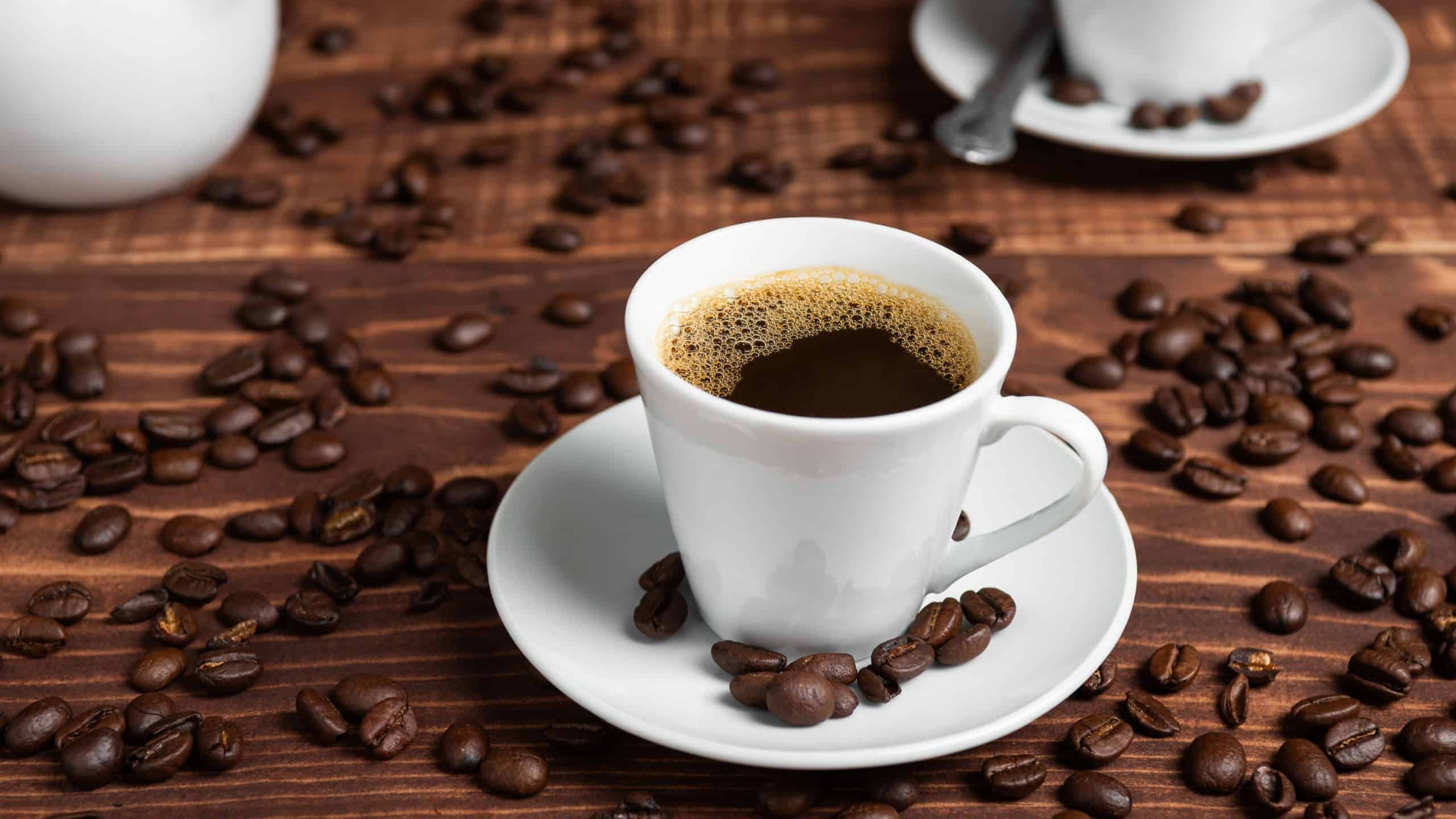 A cup of dark roasted coffee on a white saucer, surrounded by coffee beans on a brown wooden surface