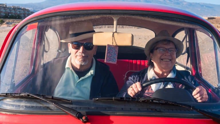 Drive an old red car for two seniors man and woman, she smiles and he looks at you. Both with hat. Alternative travel concept.