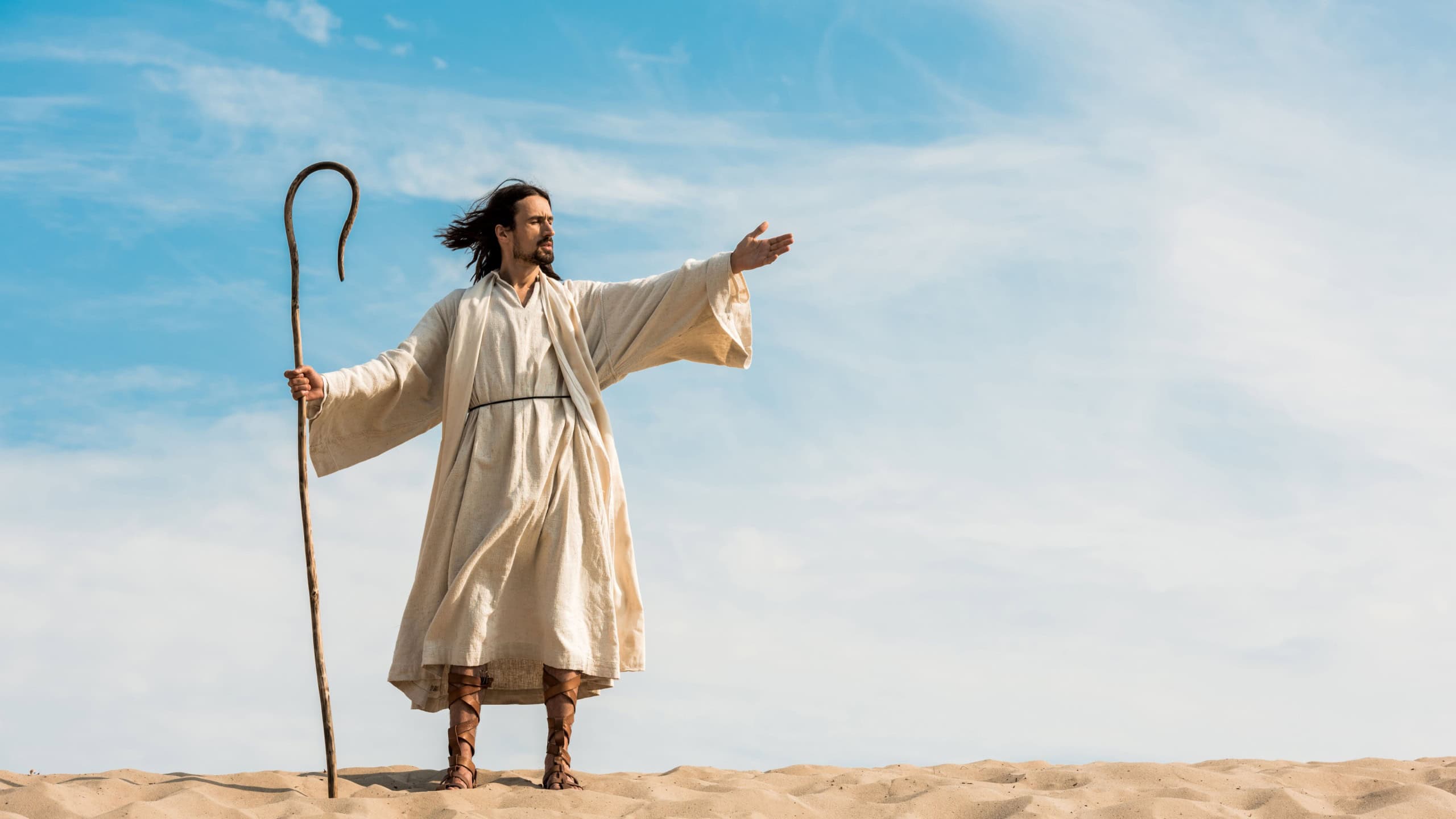 man in jesus robe holding wooden cane and gesturing against blue sky in desert