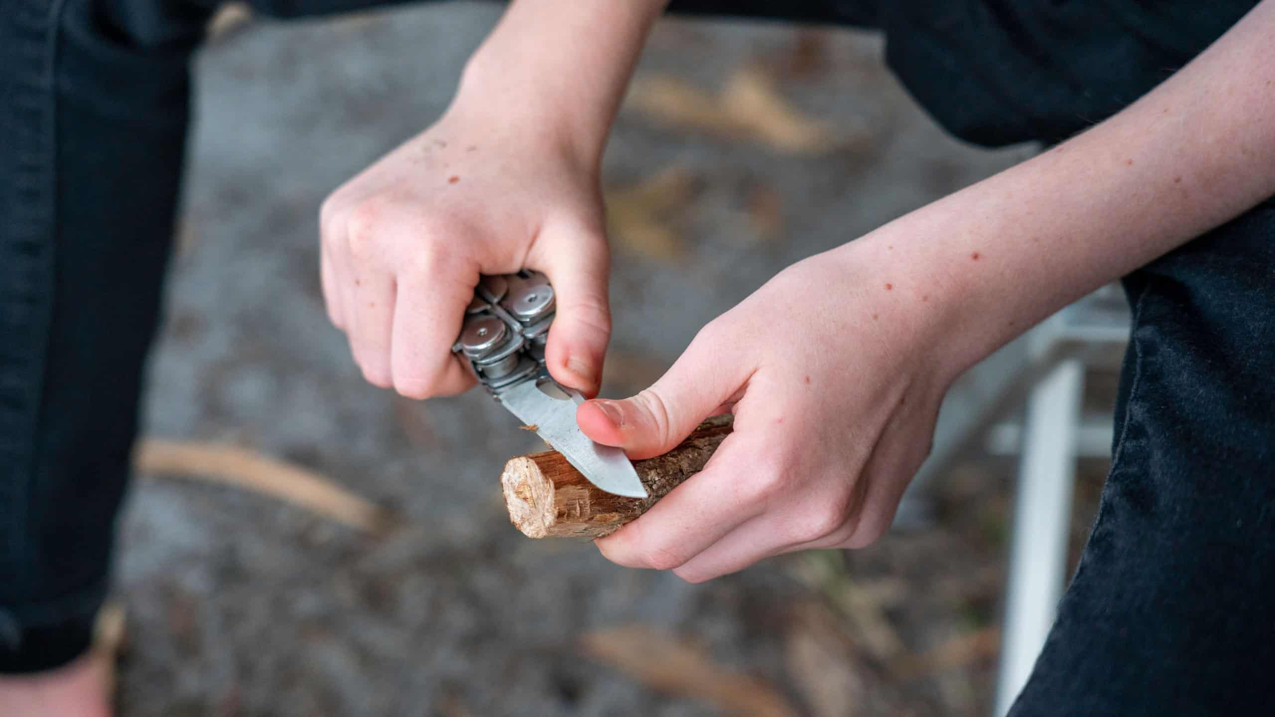 A young girl uses a knife to whittle a stick while out hiking in the wilderness.