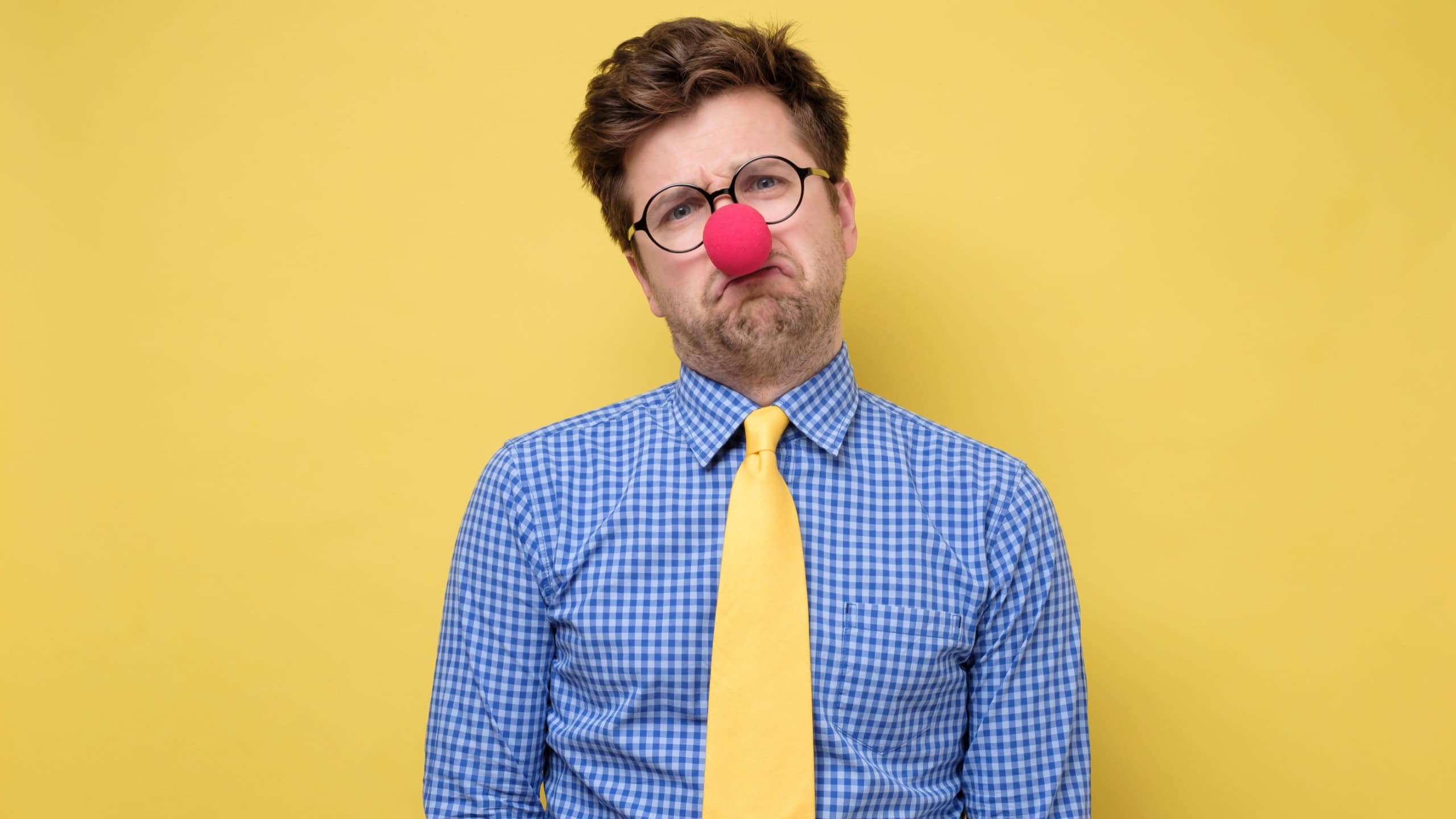Sad caucasian man wearing clown red nose on yellow wall. He is upset that nobody came to his party.