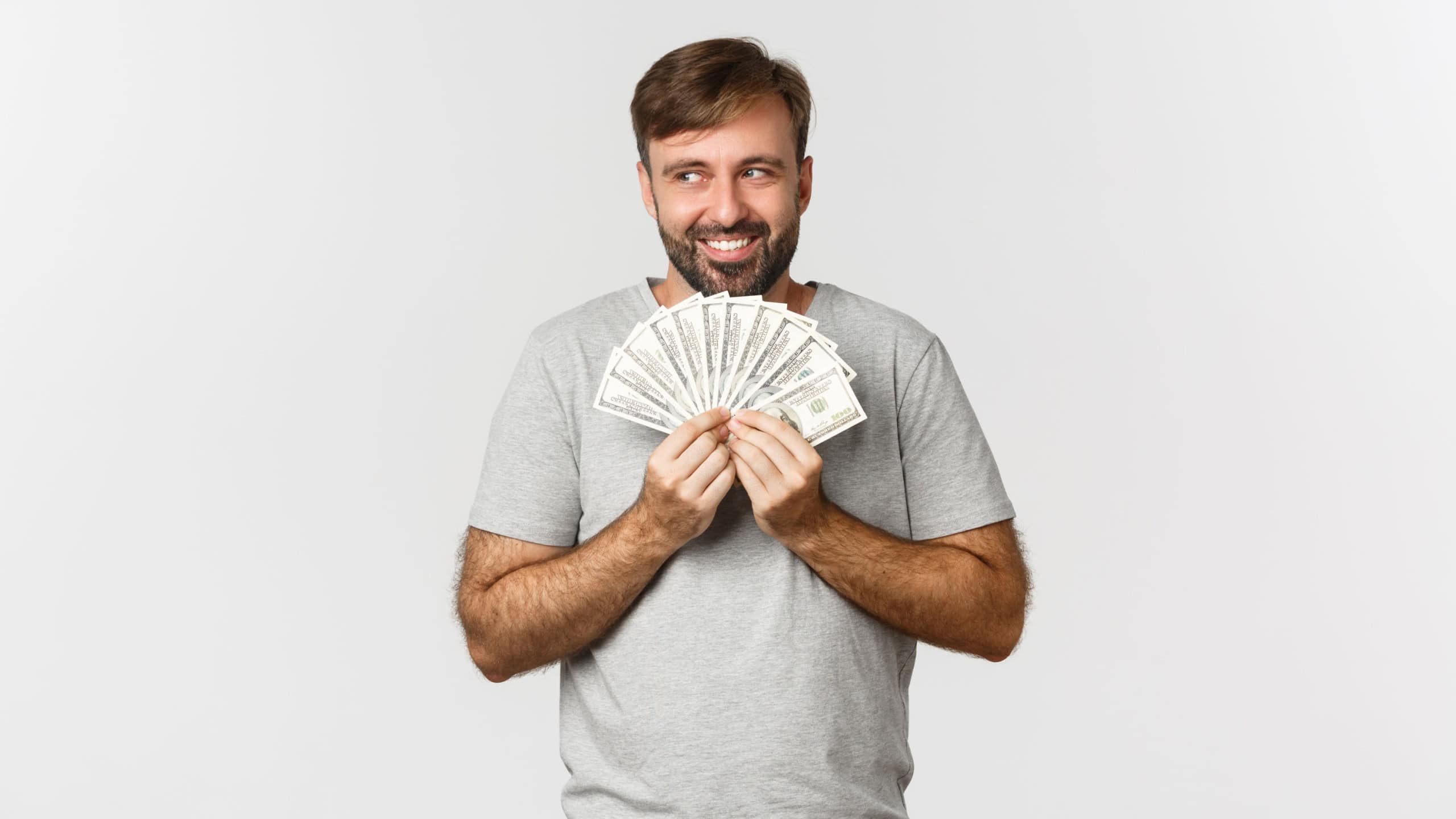 Greedy smiling greedy man with beard, thinking about shopping, holding money and looking at upper left corner, standing over white background