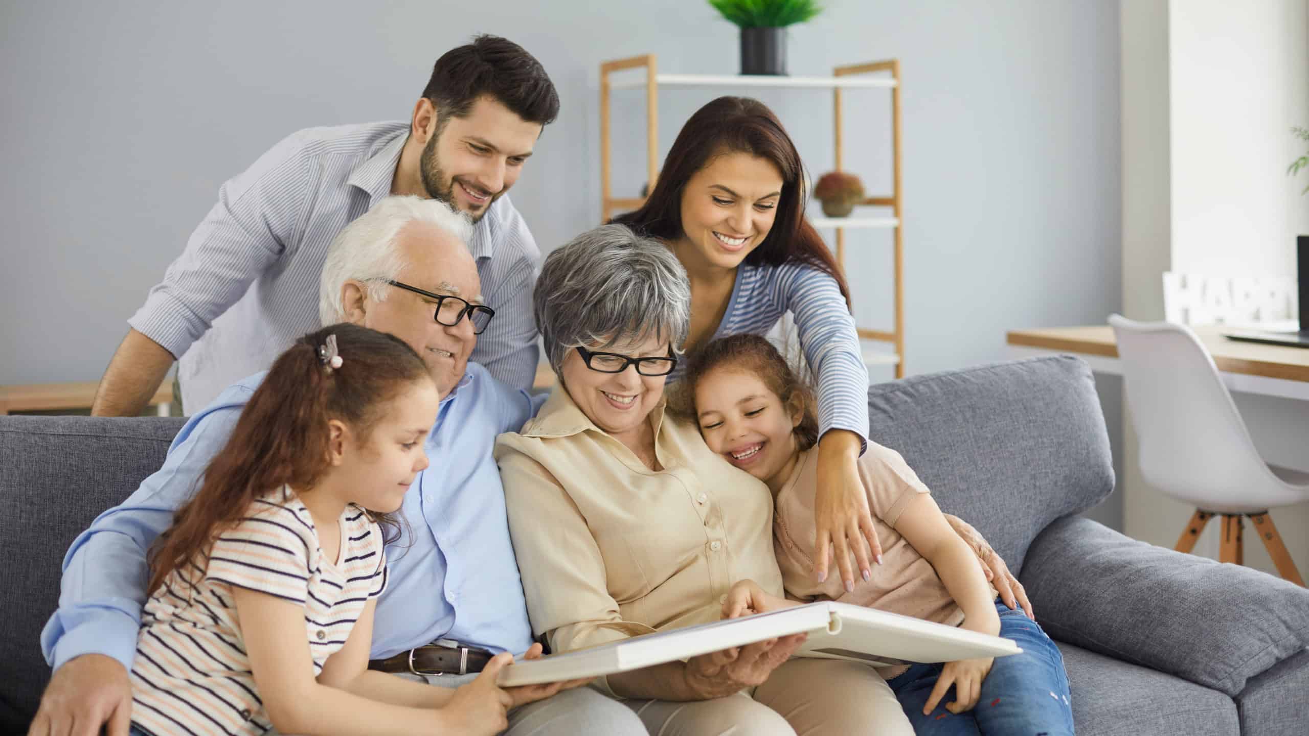 Happy big family grandparents with twin granddaughters and their parents browse the family photo album and share happy memories. Family gathered together in the living room. Family connection concept.