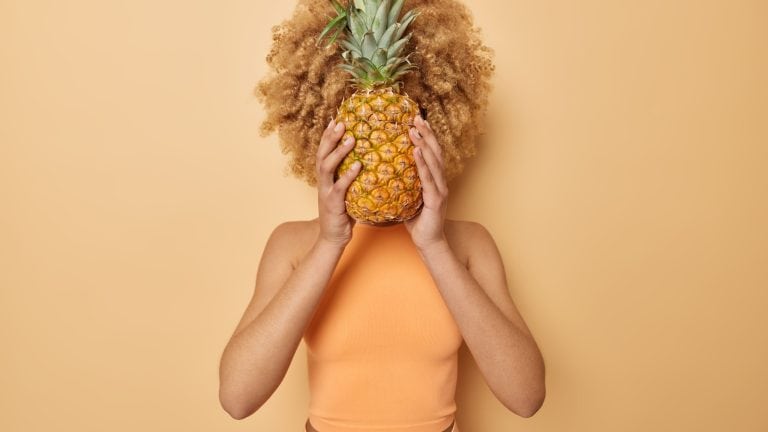 Horizontal shot of curly haired unknown woman holds pineapple over face hides herself with tropical fruit dressed in t shirt isolated over beige background. Summer time and lifestyle concept