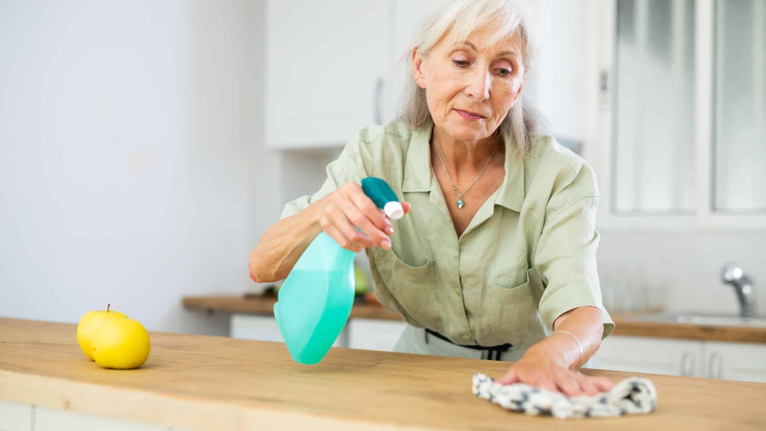 Portrait of smiling elderly woman wiping dust from kitchen surfaces with cleaning rag and detergent spray